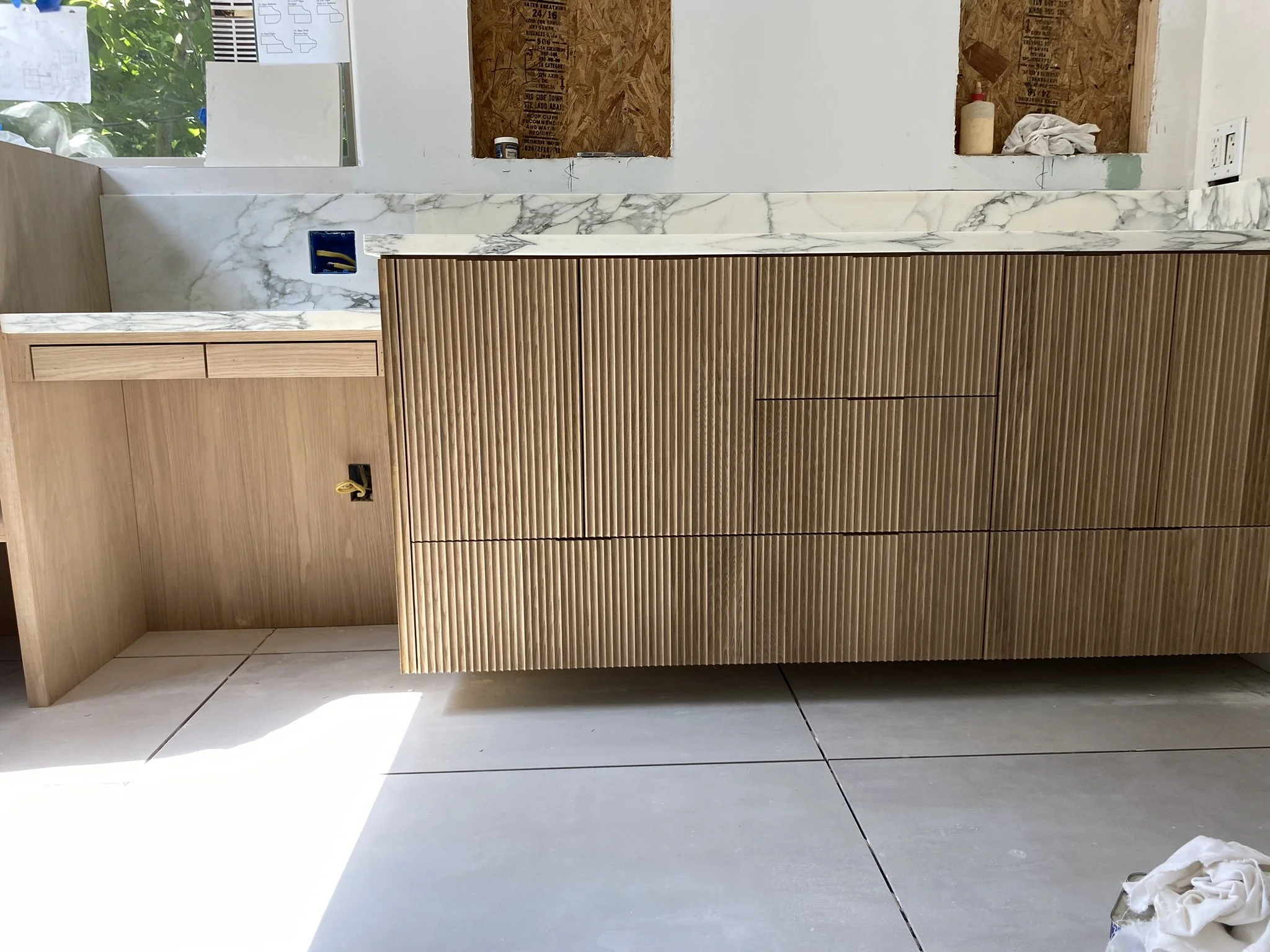 Modern bathroom vanity with ribbed wood cabinet doors, marble countertop, and tiled floor, under construction.