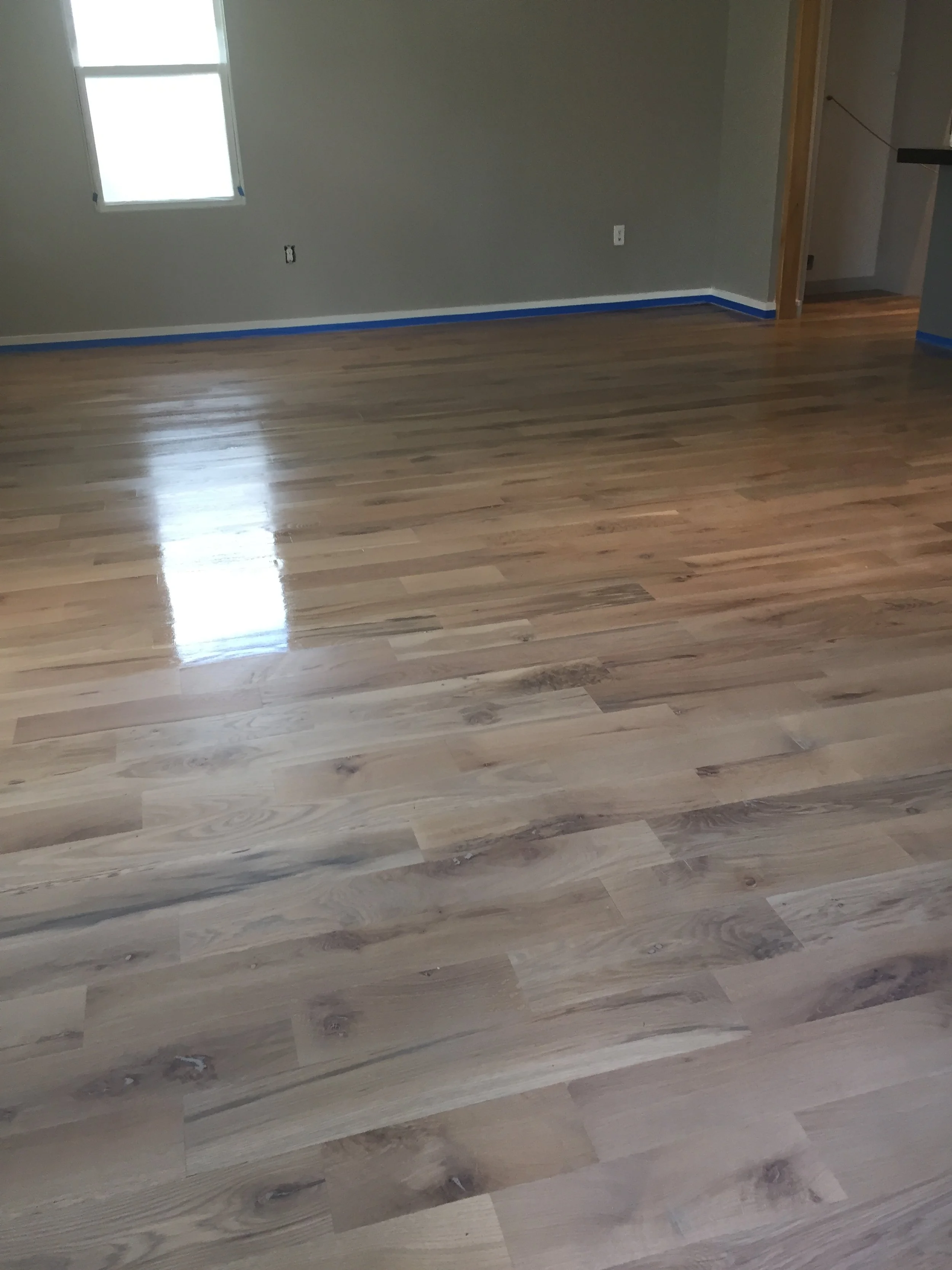 Empty room with freshly finished hardwood floor, gray walls, and a window.