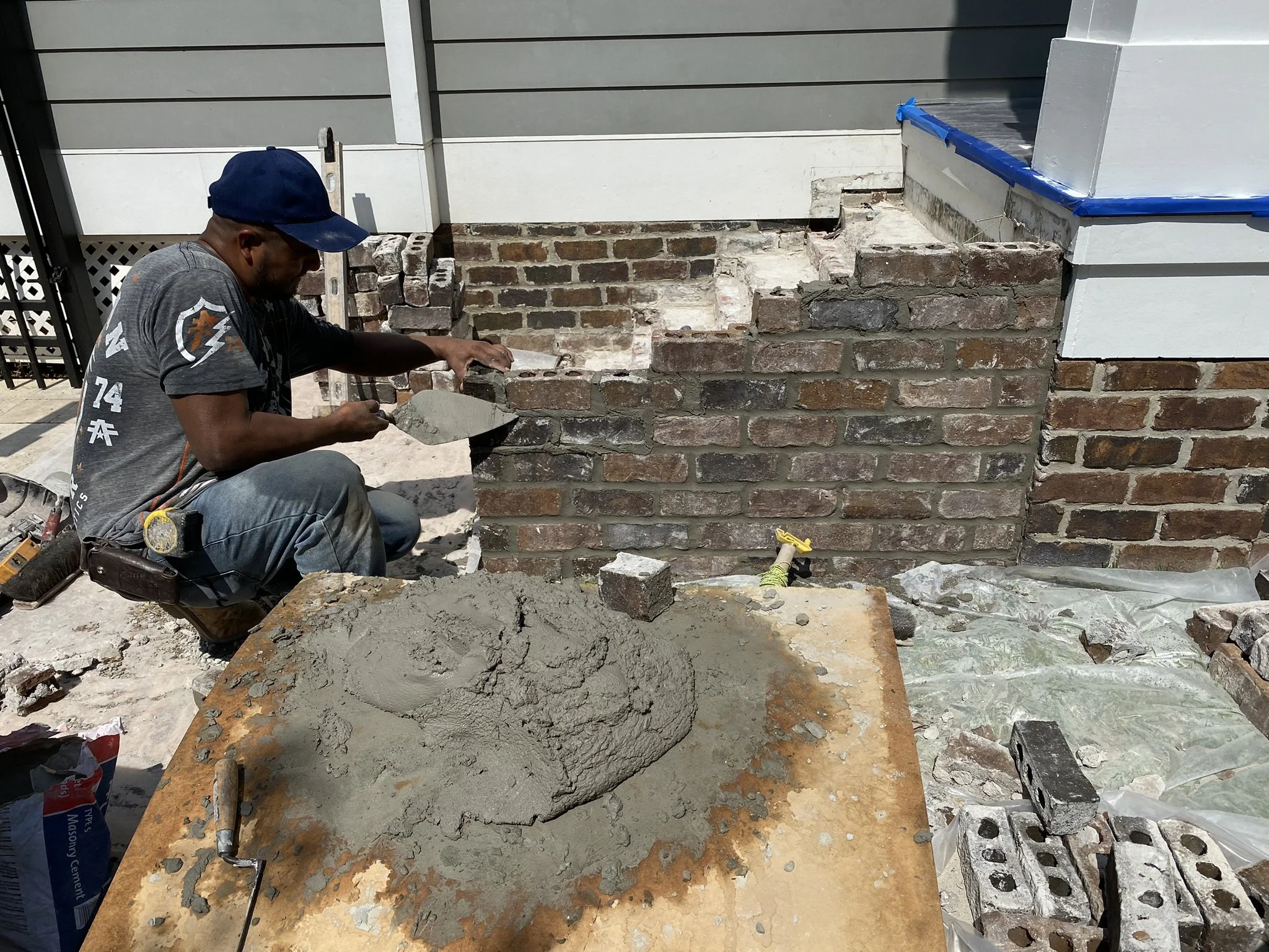 J. Luis Construction Inc worker is applying mortar to bricks, repairing or building a brick wall. rowel and has a mix of concrete on a wooden board beside them. 