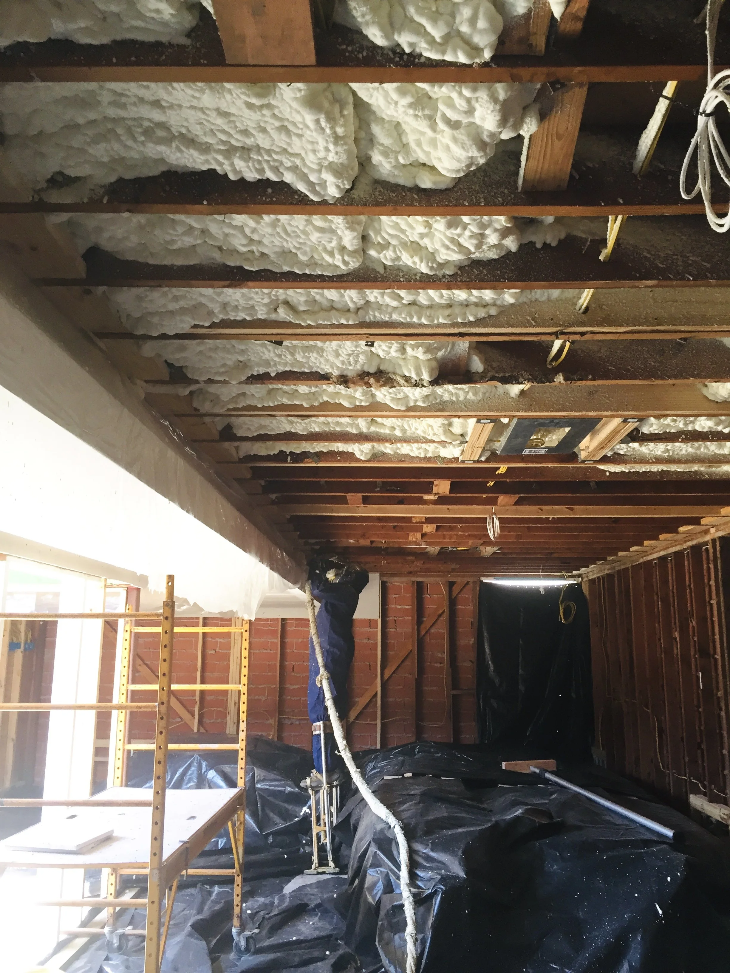 Construction site with exposed ceiling beams and spray foam insulation installation. Worker on a ladder applying insulation, with scaffolding and protective coverings on floor.