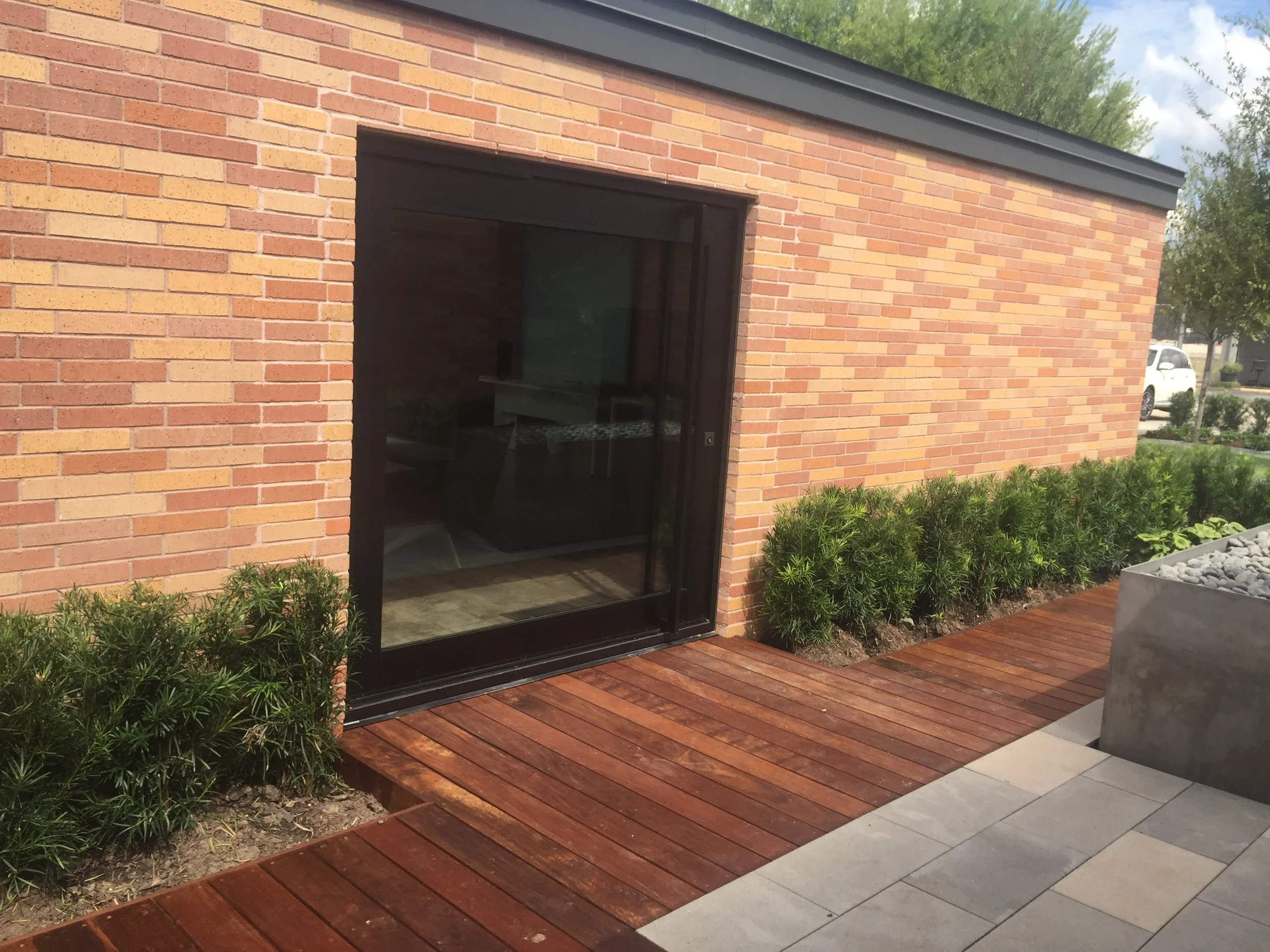 Brick building with sliding glass door, wooden deck, and small shrubs next to the wall.
