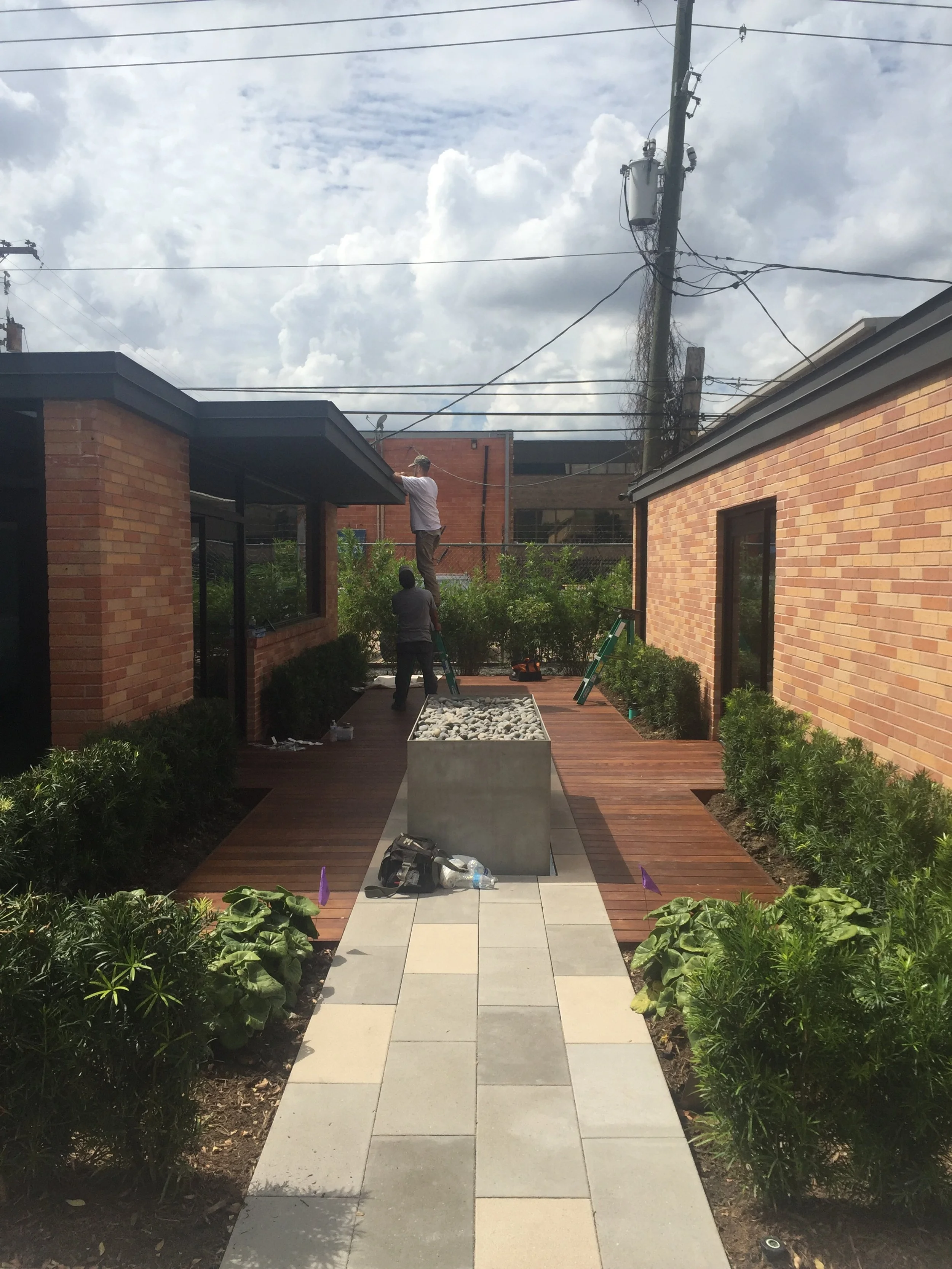 Outdoor patio area with brick buildings, wooden deck, planter with rocks, two people on ladders working, and surrounding greenery.