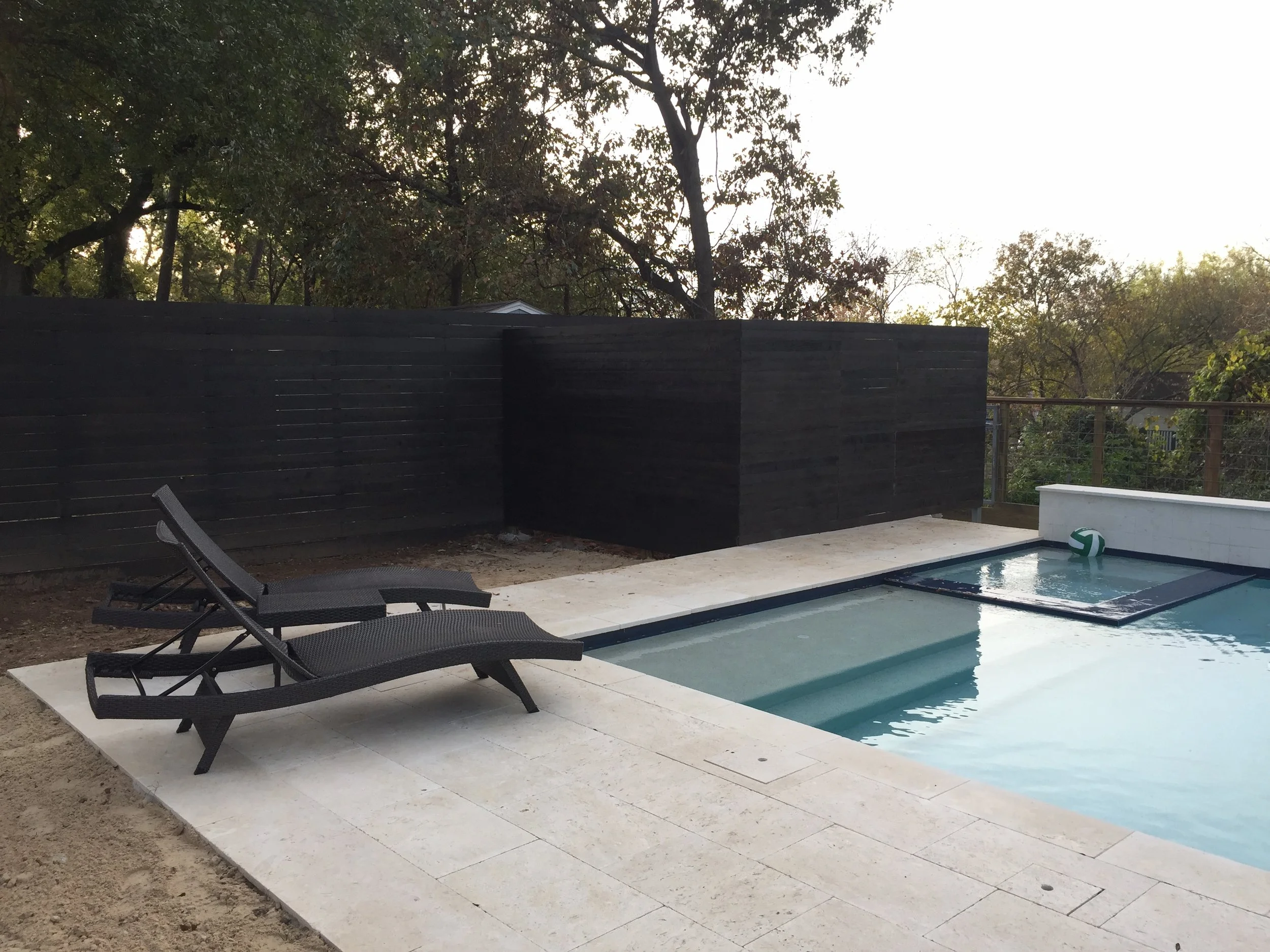 A backyard swimming pool with two dark wicker lounge chairs on a light stone patio. The pool has a small square section with a volleyball floating in it. A wooden privacy fence surrounds the area, with trees in the background.