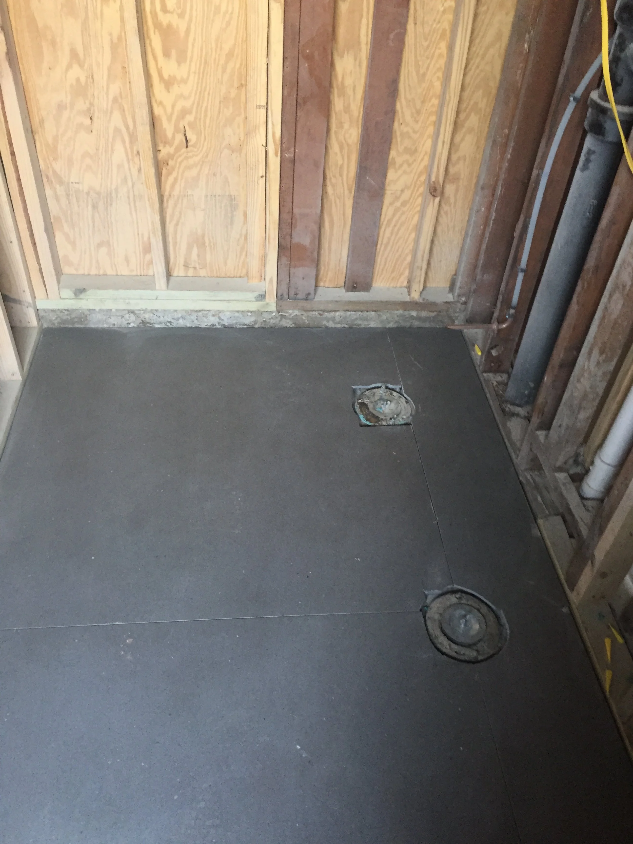 Unfinished bathroom construction with plywood walls and a dark cement board floor, featuring a drain opening.