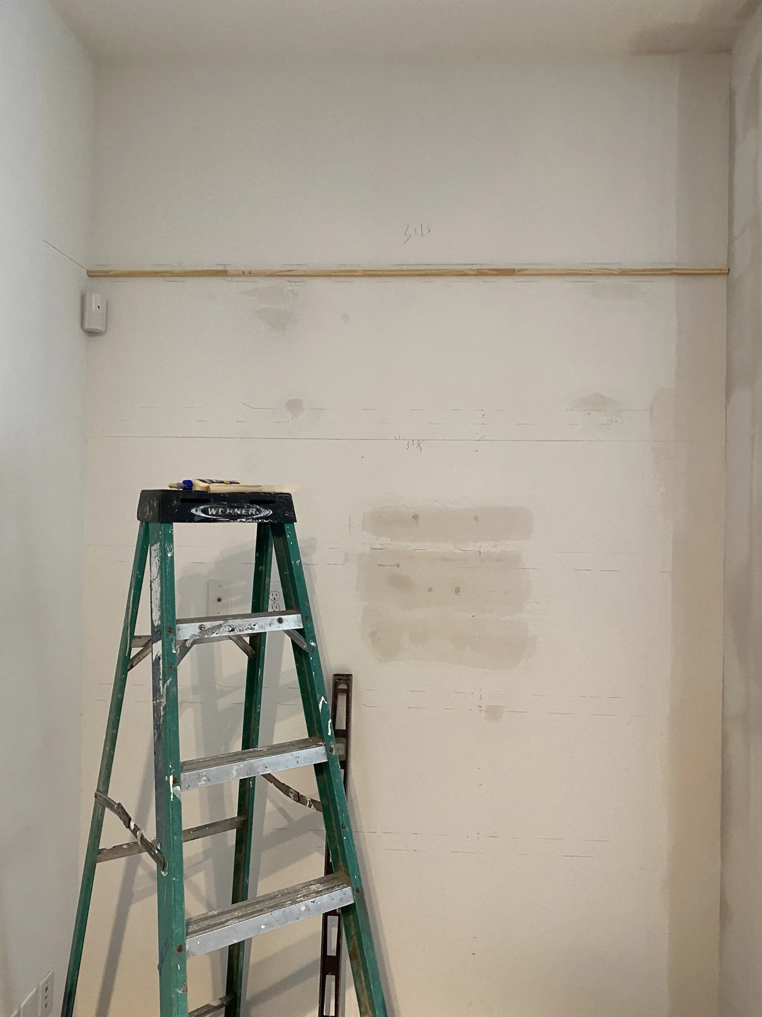 A green step ladder leaning against an unfinished wall, with patched areas and a wooden beam attached horizontally. There's a light switch and a level resting on the ladder.