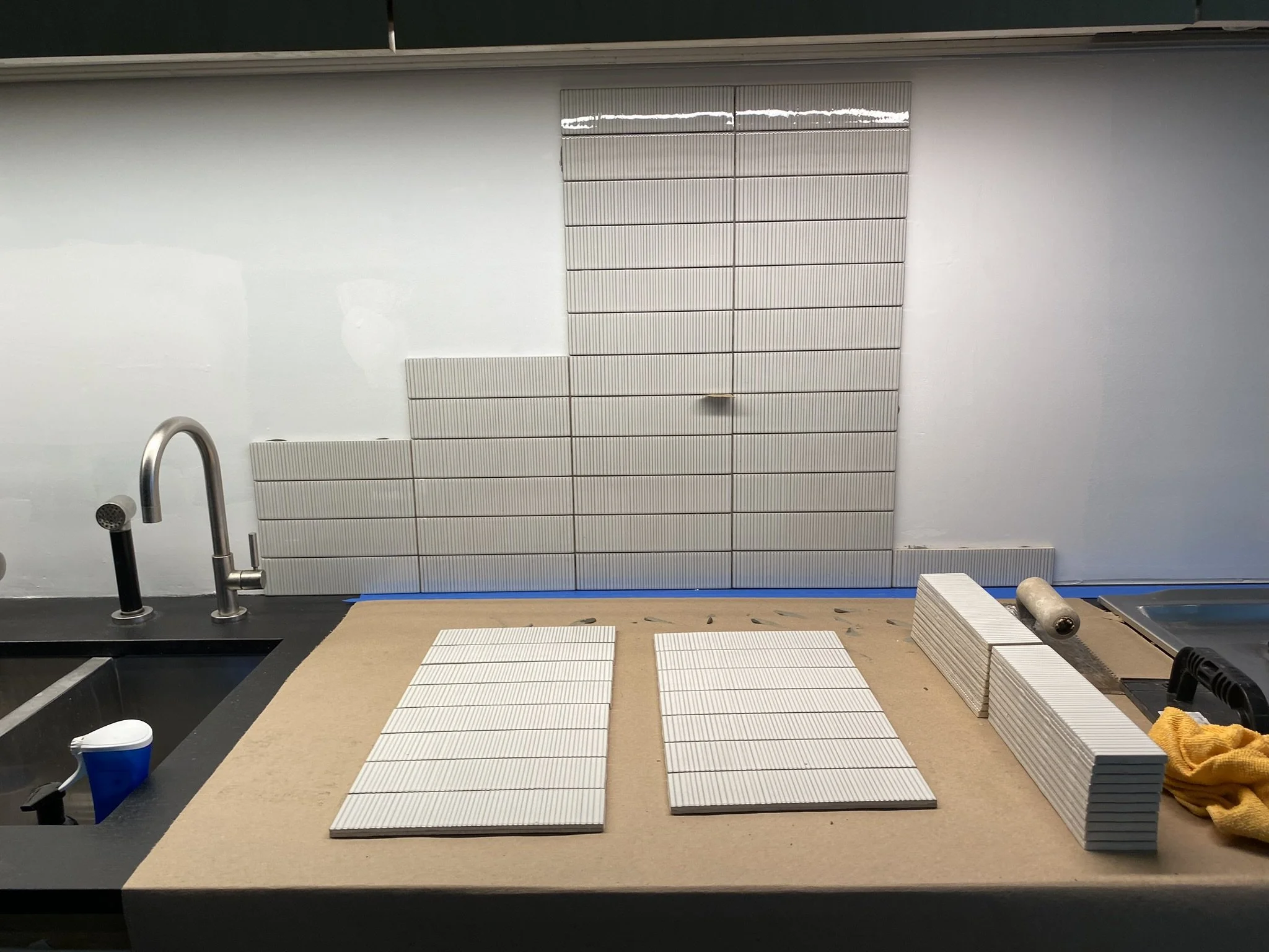 Partially tiled kitchen backsplash with white rectangular tiles, stainless steel faucet, and stacked tiles on a countertop.