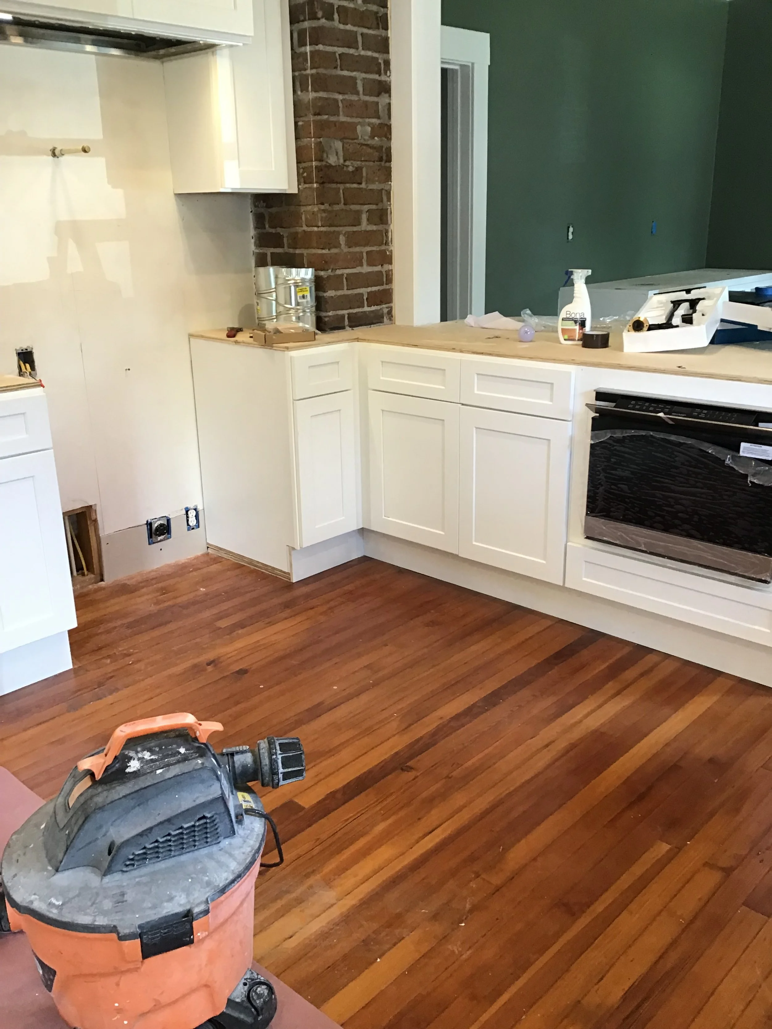 Kitchen under renovation with white cabinets, exposed brick wall, hardwood floors, and a vacuum cleaner on the floor.