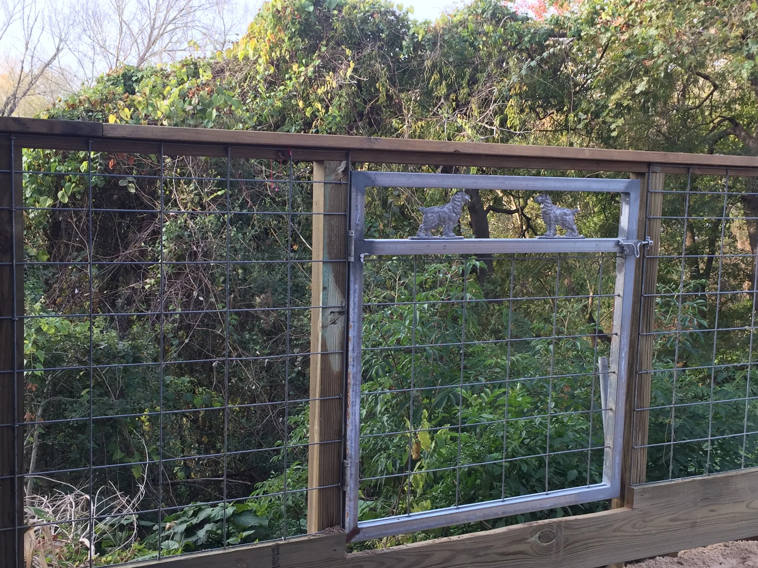 A wire mesh fence with wooden posts and a metal gate featuring dog silhouettes, surrounded by lush green foliage.
