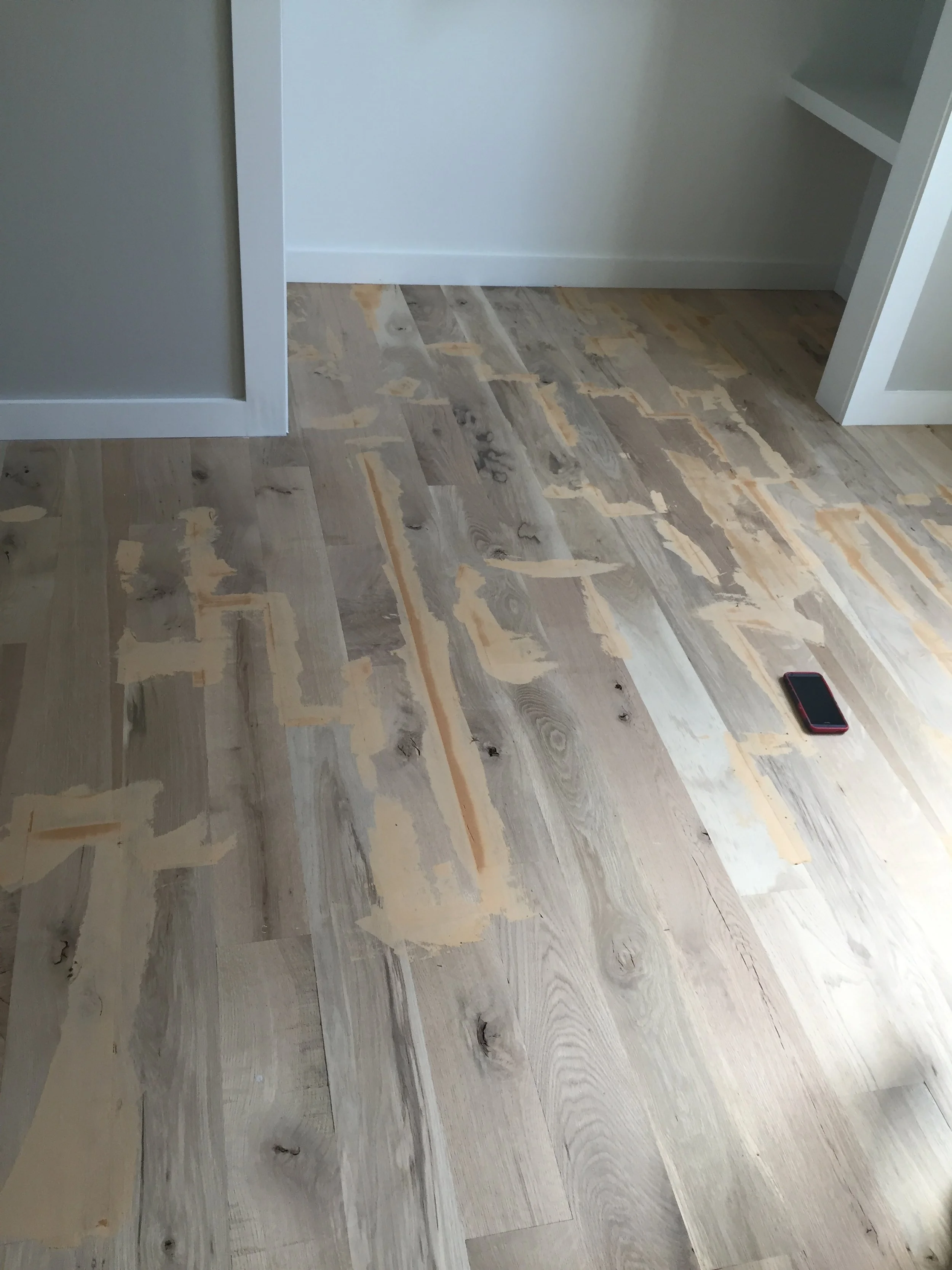 Unfinished hardwood floor with patches of wood filler and black smartphone on the surface, next to a white wall and shelves.
