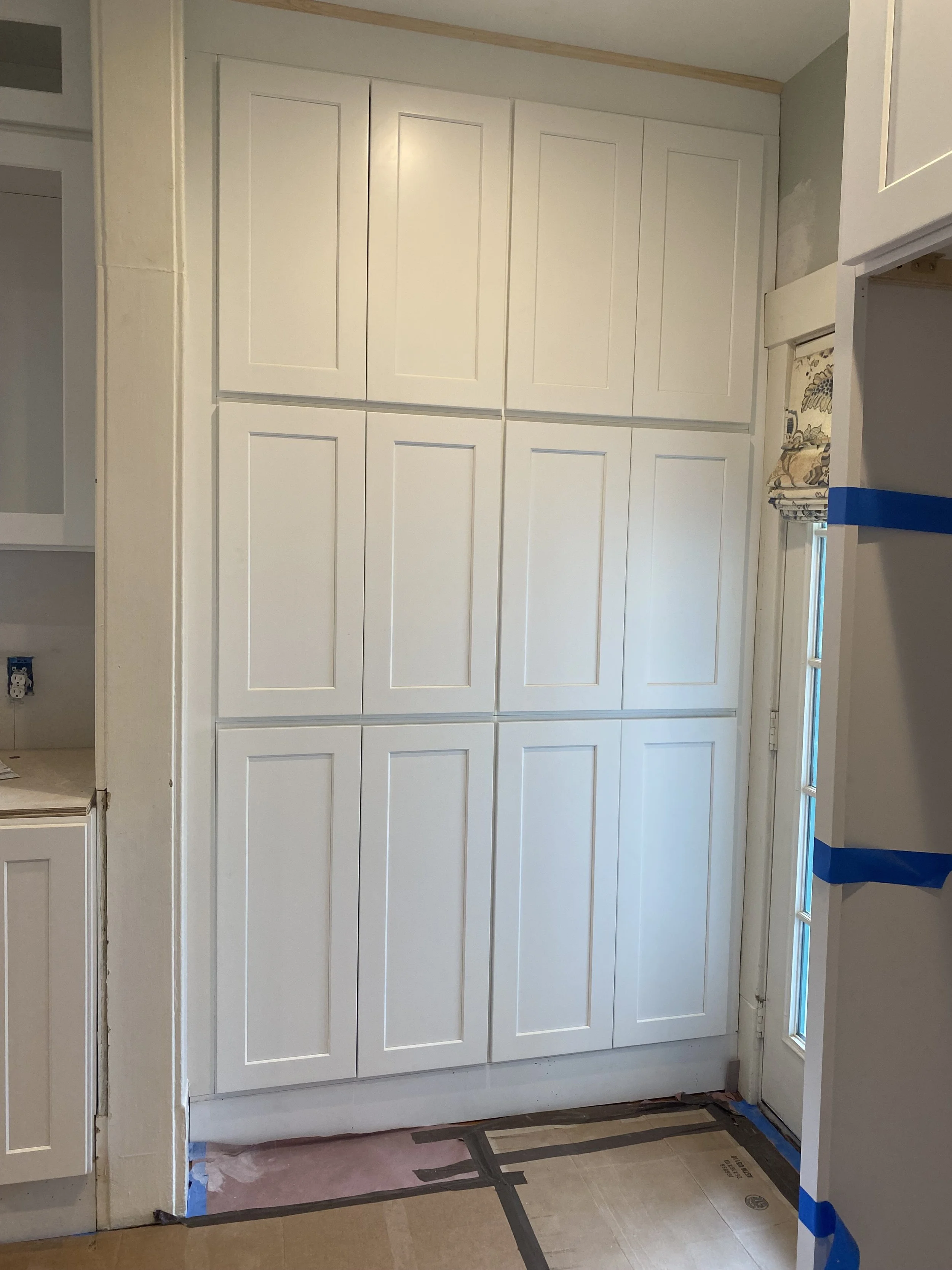 White kitchen cabinets with a raised panel design, adjacent to a door with a decorative curtain, and a taped floor indicating ongoing renovation.