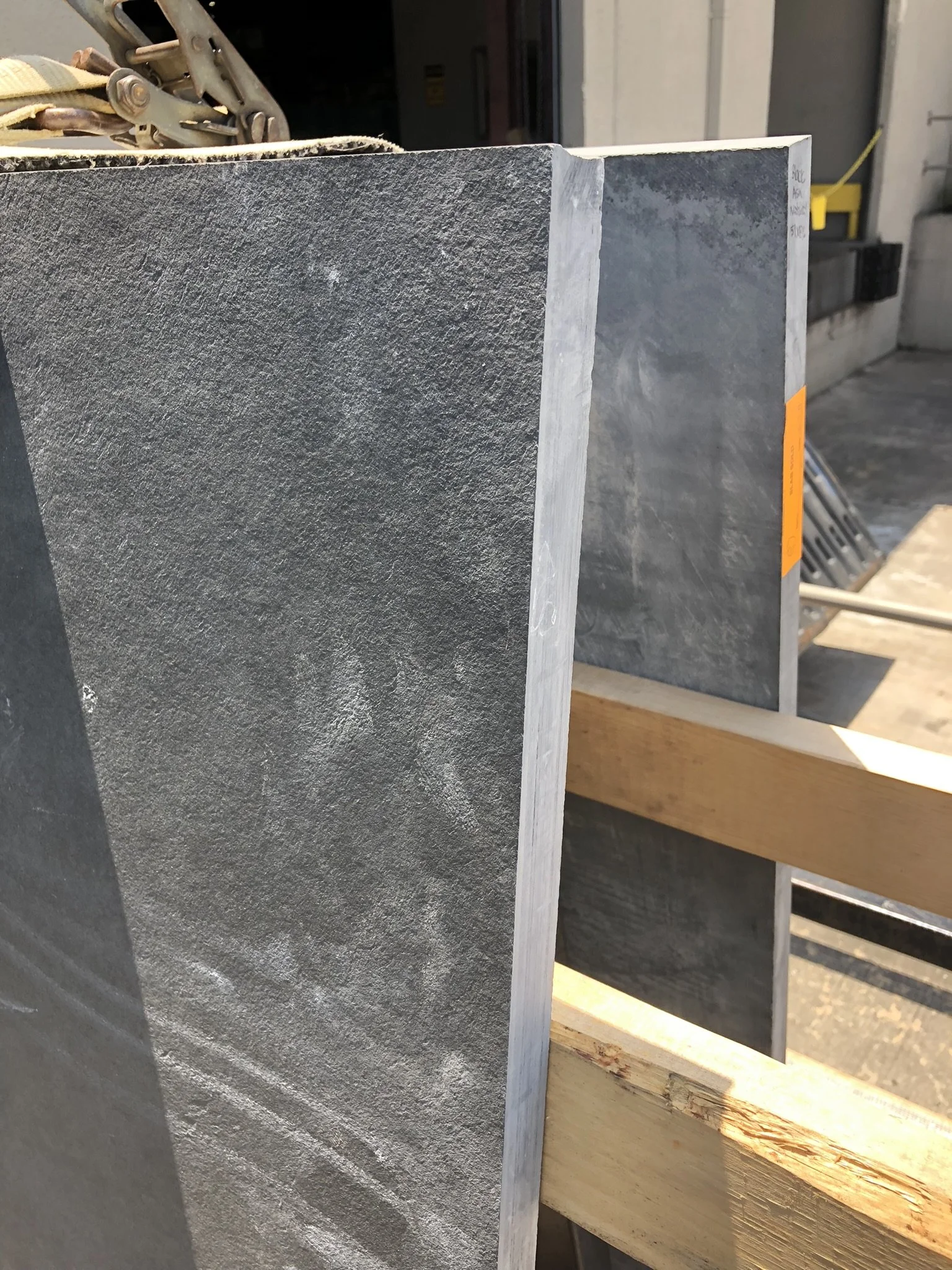 Close-up of stacked gray stone slabs with a textured surface, secured with straps on a wooden frame, in an outdoor setting.