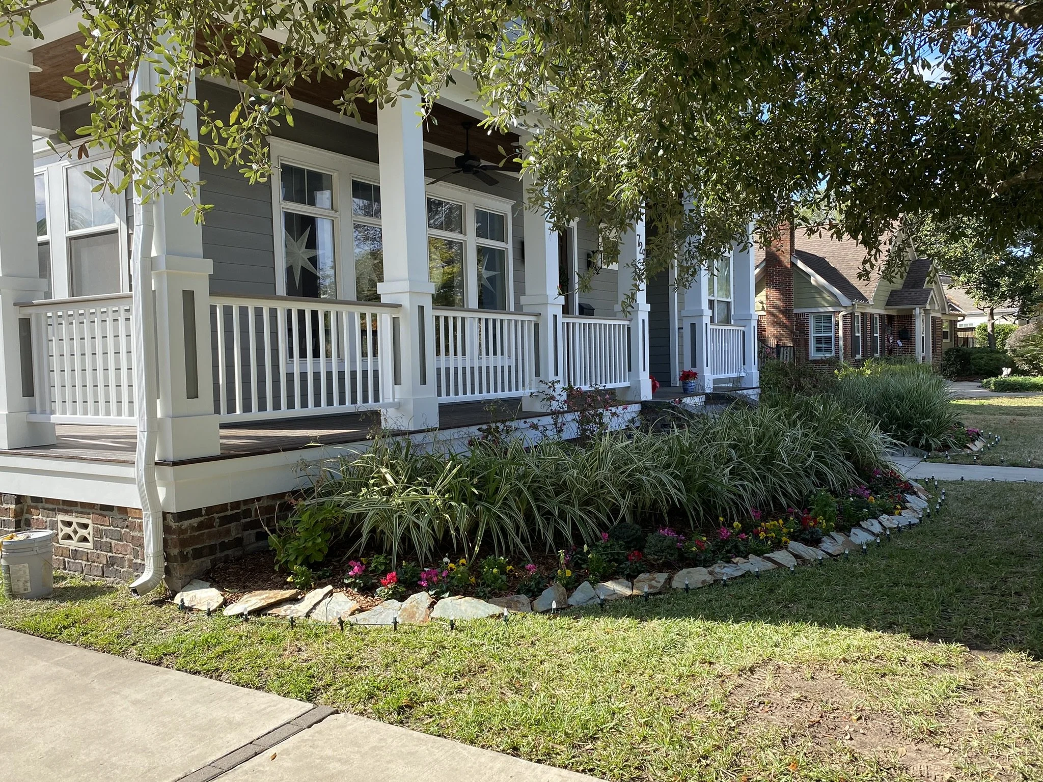 Suburban house with a wide porch, white railing, and a landscaped garden with plants and flowers, situated in a grassy front yard.