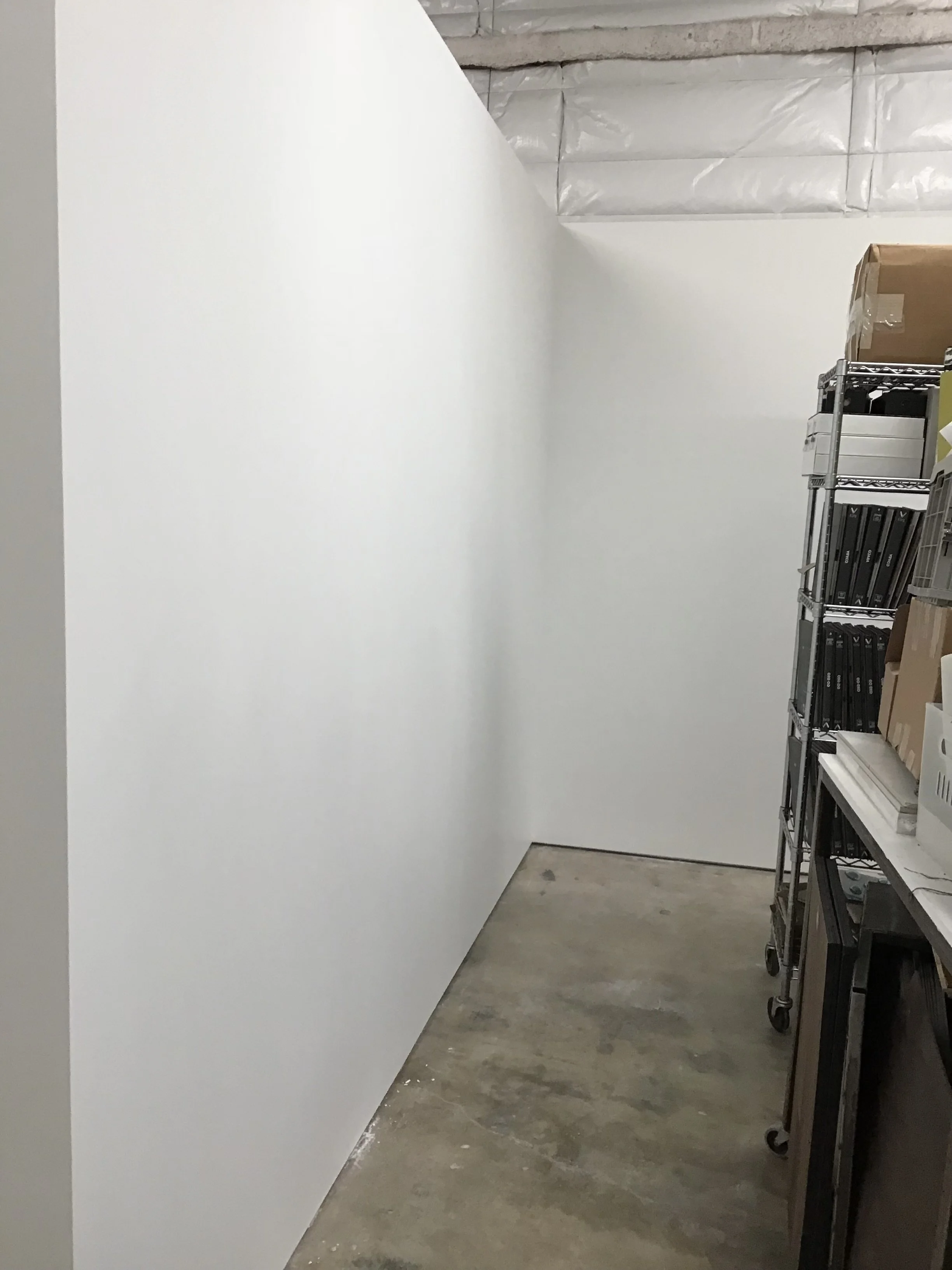 Narrow storage area with white walls, concrete floor, metal shelving holding boxes and binders, industrial ceiling.