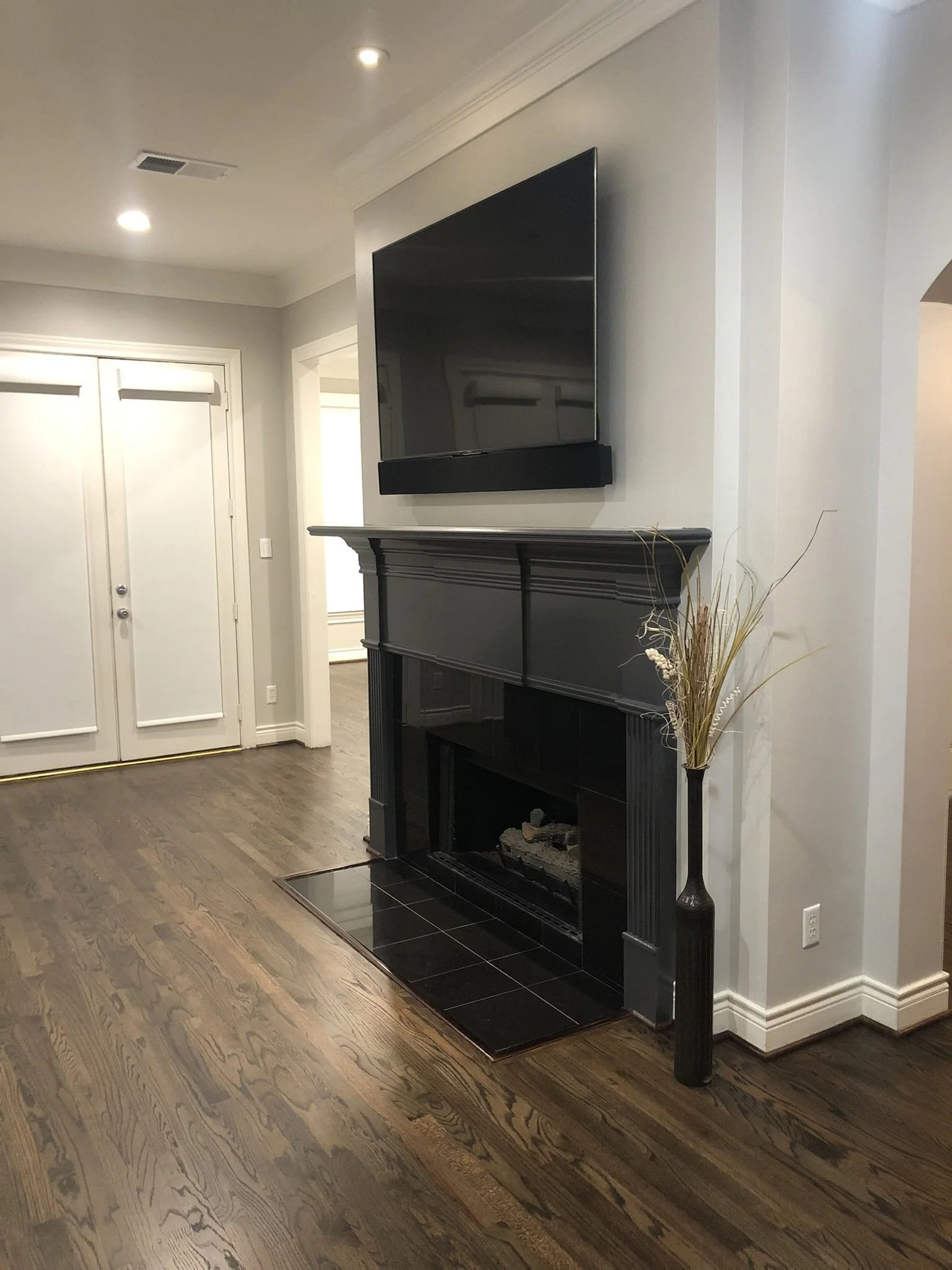 Modern living room with a wall-mounted TV above a black fireplace, dark hardwood floors, and a decorative vase with dried branches.