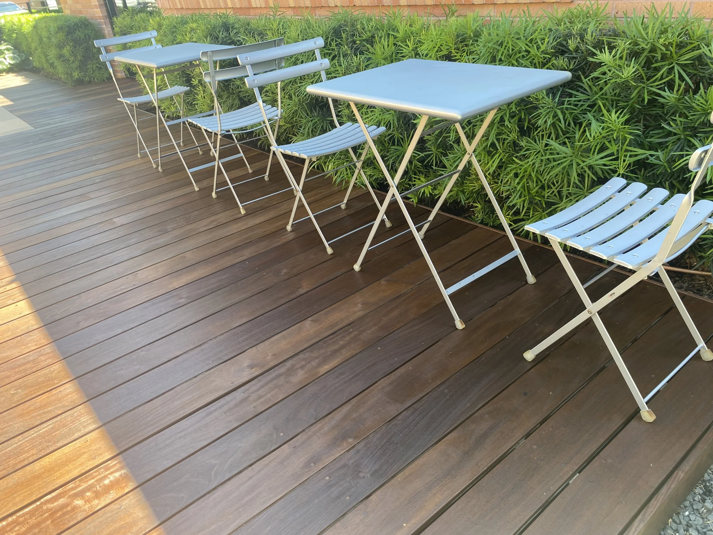 Outdoor patio with wooden decking and metal folding tables and chairs next to green shrubs.