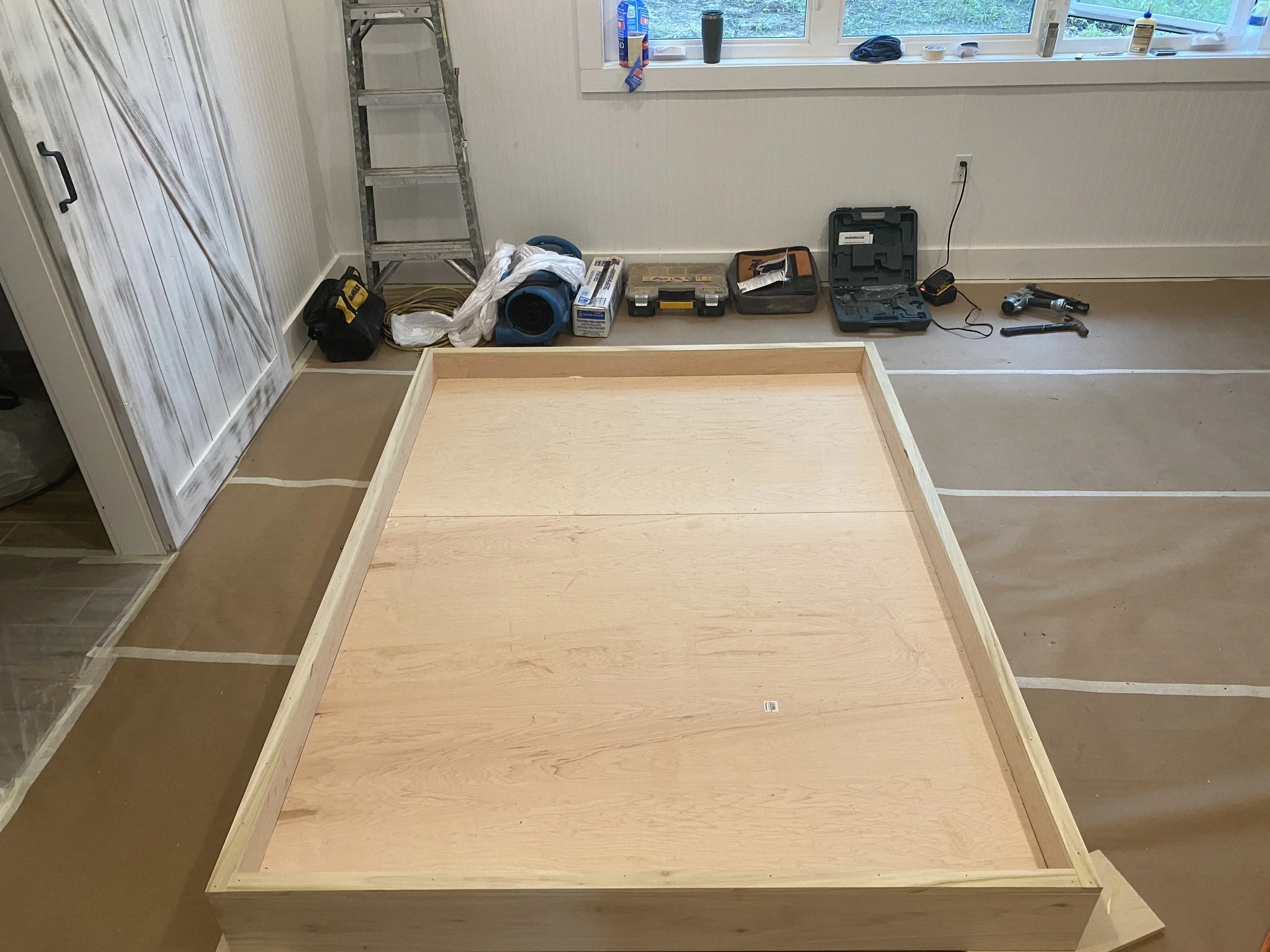 Room under construction with a wooden frame on the floor, tools and ladder against the wall, and a window with items on the sill.