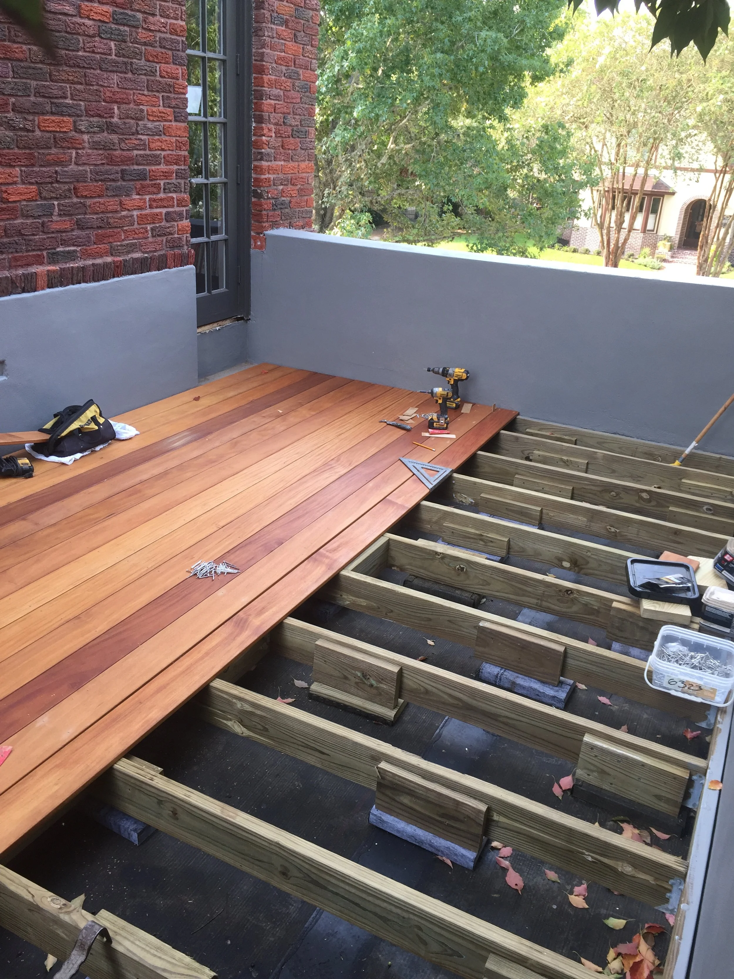 A partially constructed outdoor deck with wooden boards being installed on a frame. Tools like a drill and measuring square are visible on the wooden deck. The background shows a brick wall and leafy green trees.