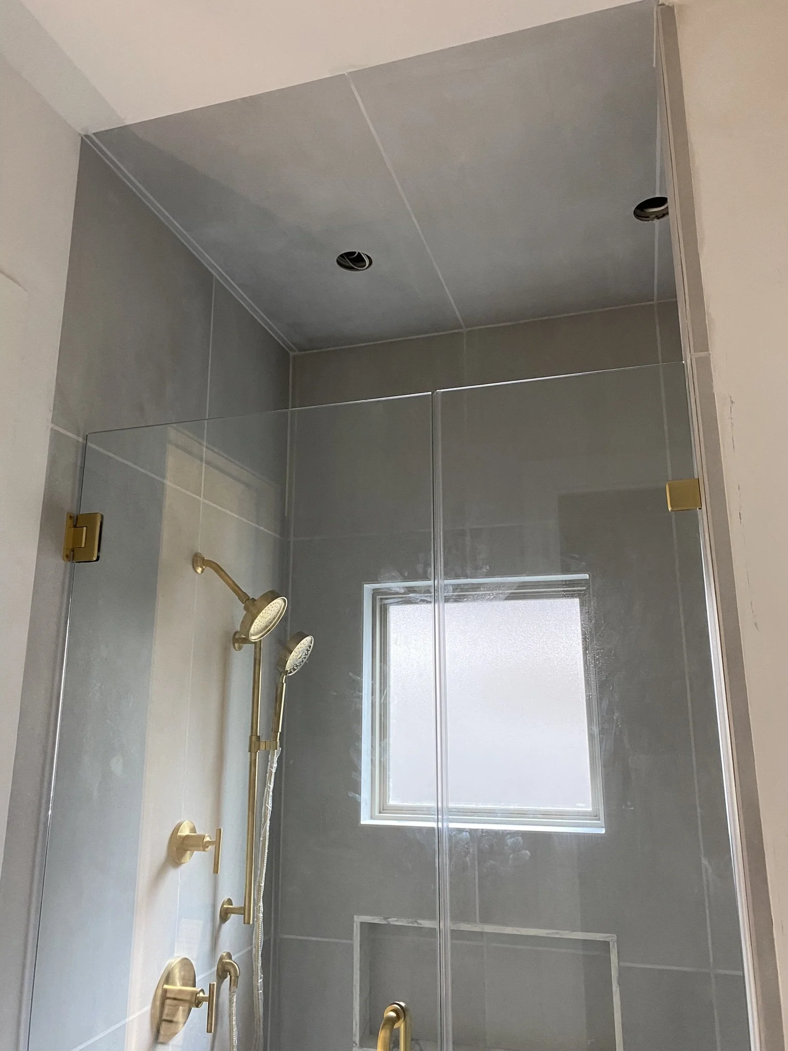 Modern shower with glass doors and gold fixtures, including dual shower heads and controls. A frosted window is on the wall.