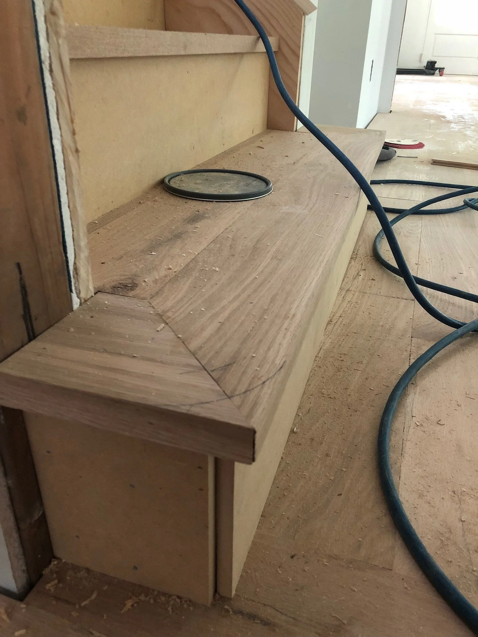 Unfinished wooden staircase with construction materials, including a blue cable, on the floor. A round lid or cover is placed on the step.