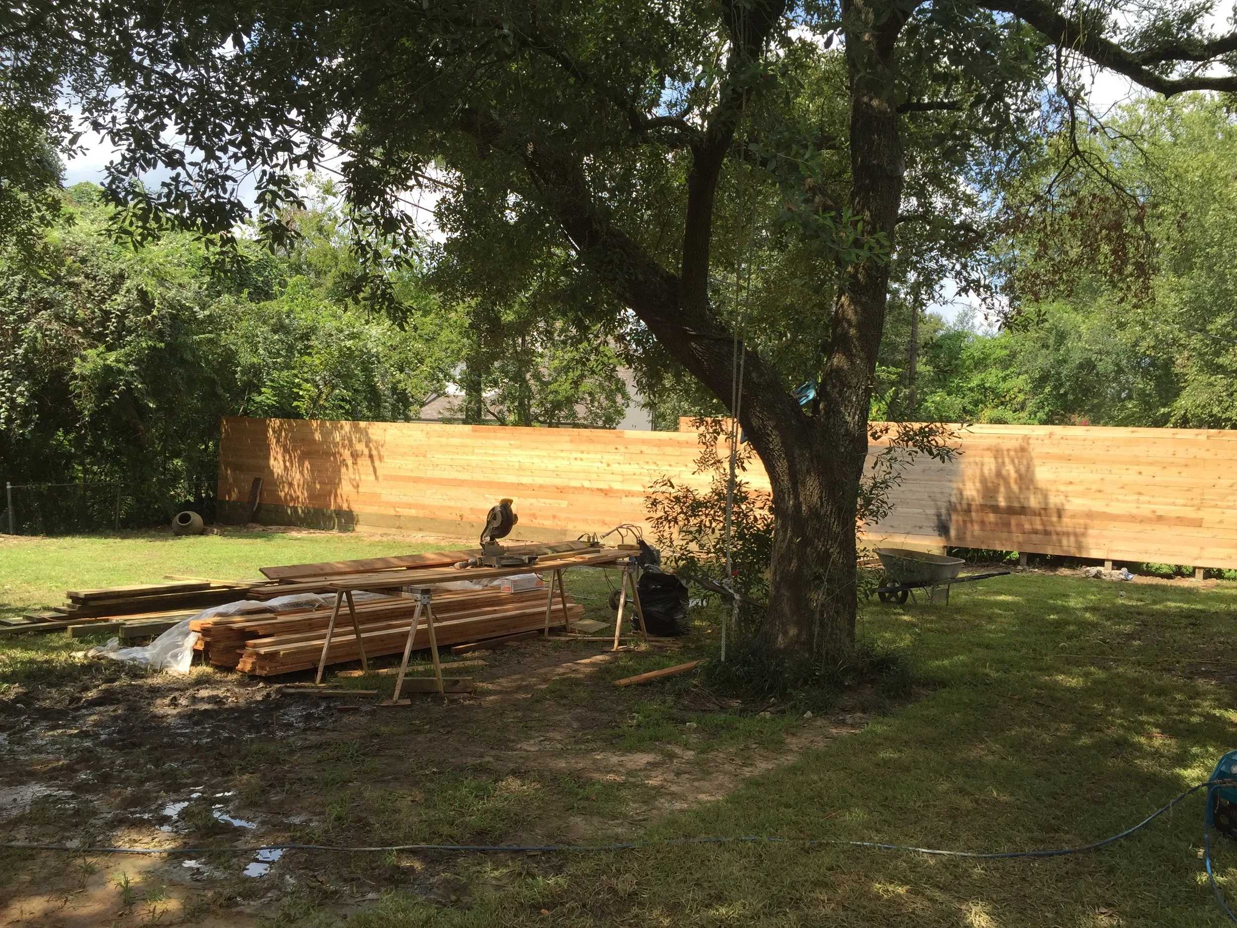Wooden fence under construction in a backyard with trees, lumber, and construction tools scattered.