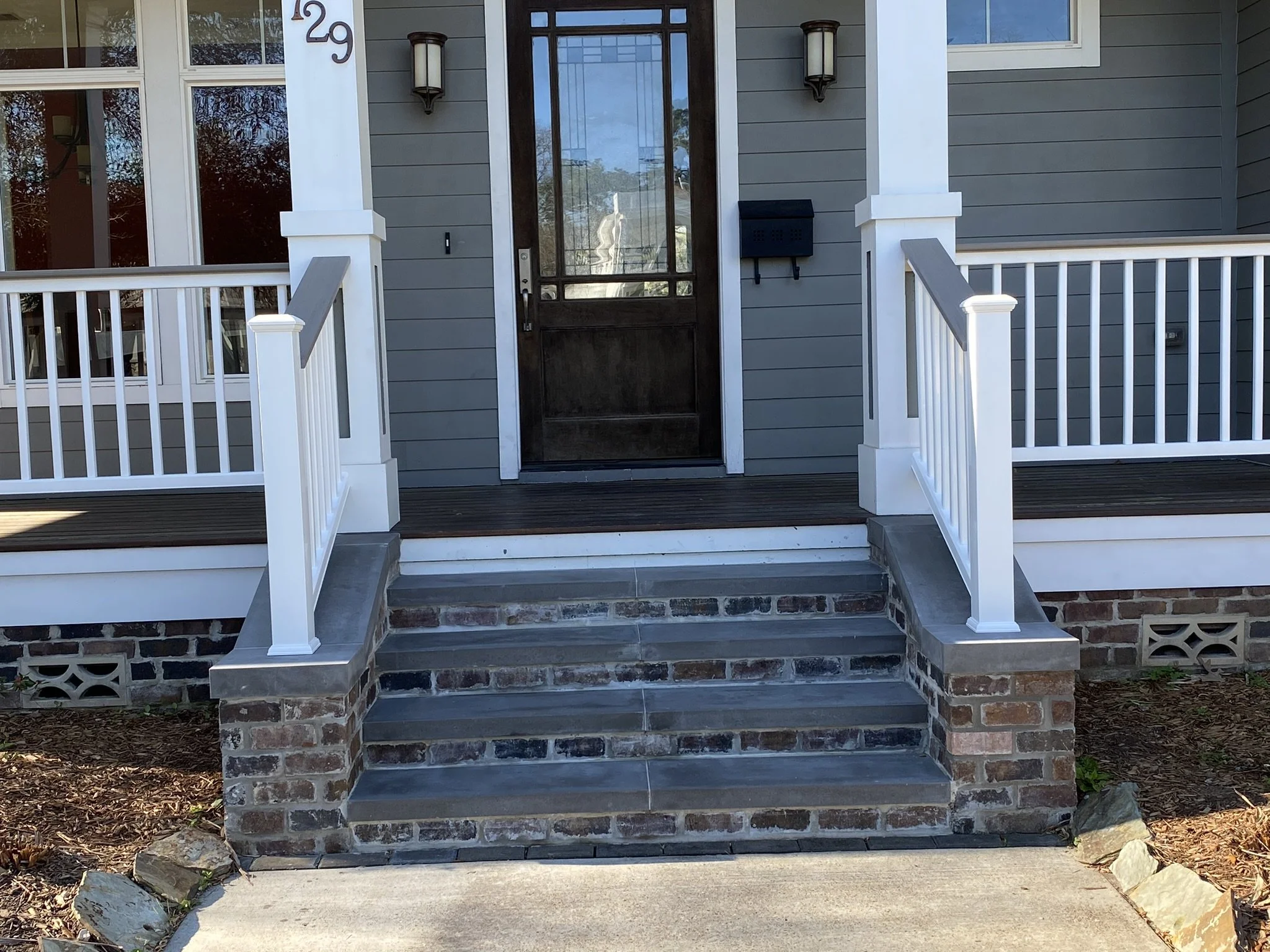 Front porch of a house with gray siding, brick steps, and white railings leading to a wooden door.