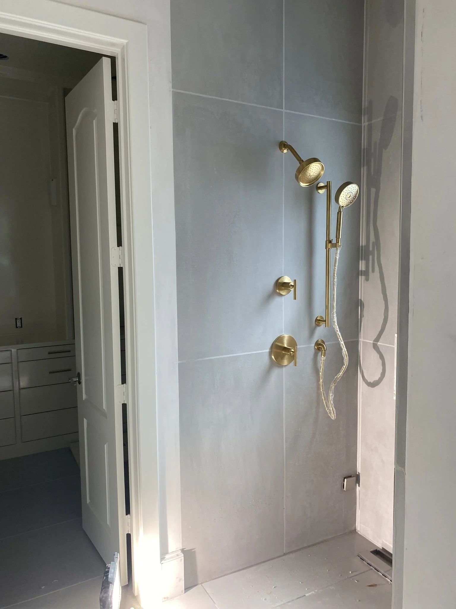 Modern shower area with gold fixtures and gray tiles next to an open doorway leading to a white cabinet.