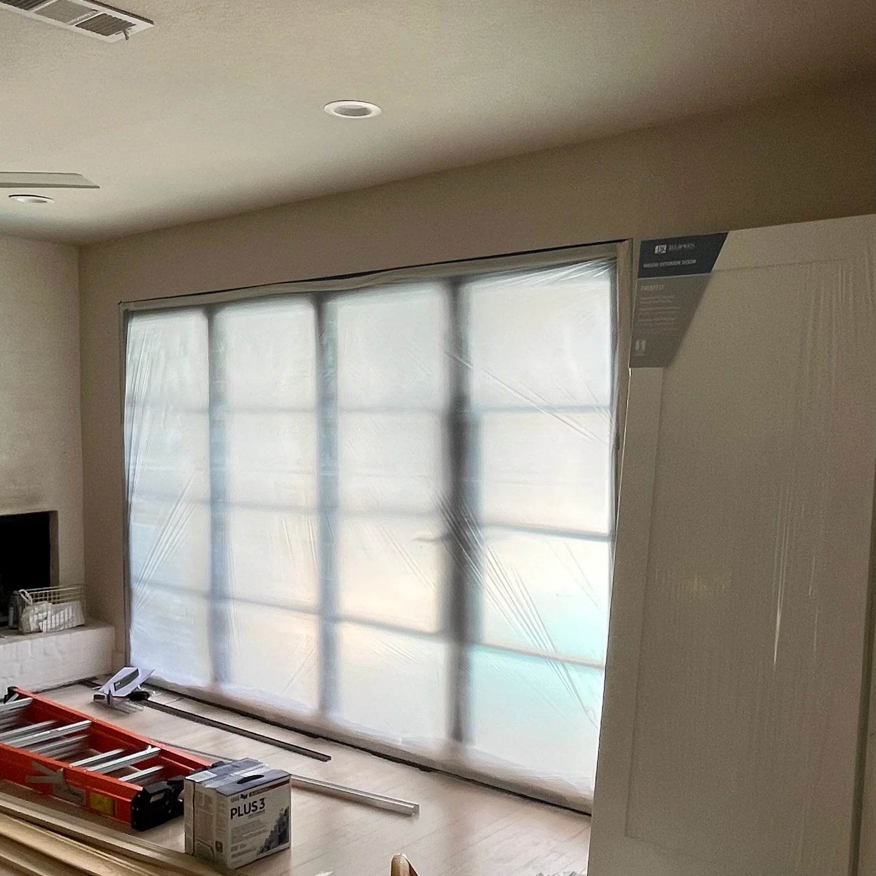 Room under renovation with large window covered in plastic, red ladder, drywall, and construction materials visible.