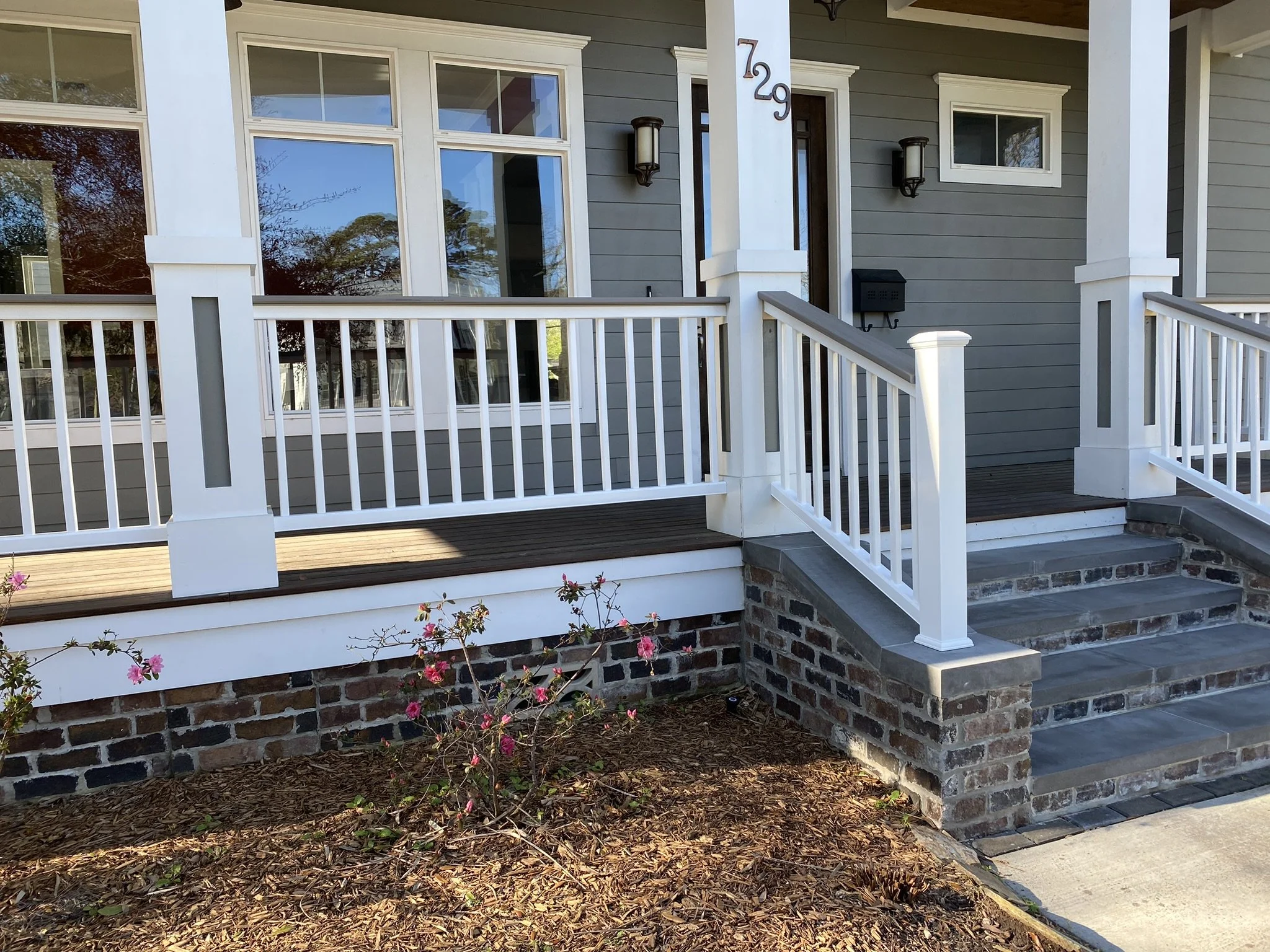 Front porch of a house with a wooden deck and white railing, gray siding, steps made of stone bricks, and some small flowering plants in the garden bed below.