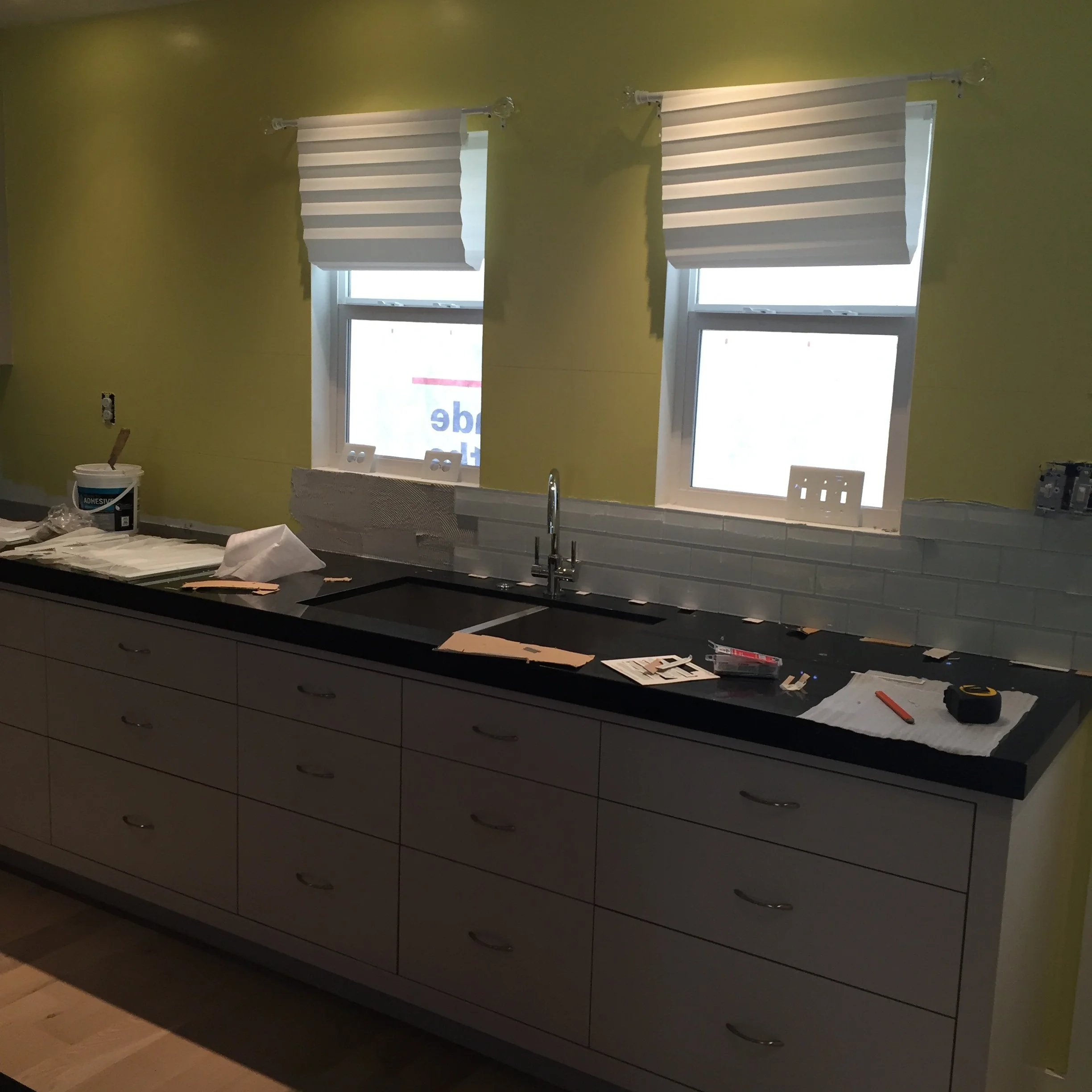 Kitchen under renovation with white drawers, dark countertop, yellow walls, and white subway tile backsplash partially installed. Two windows with white blinds above the sink. Tools and materials on the counter.