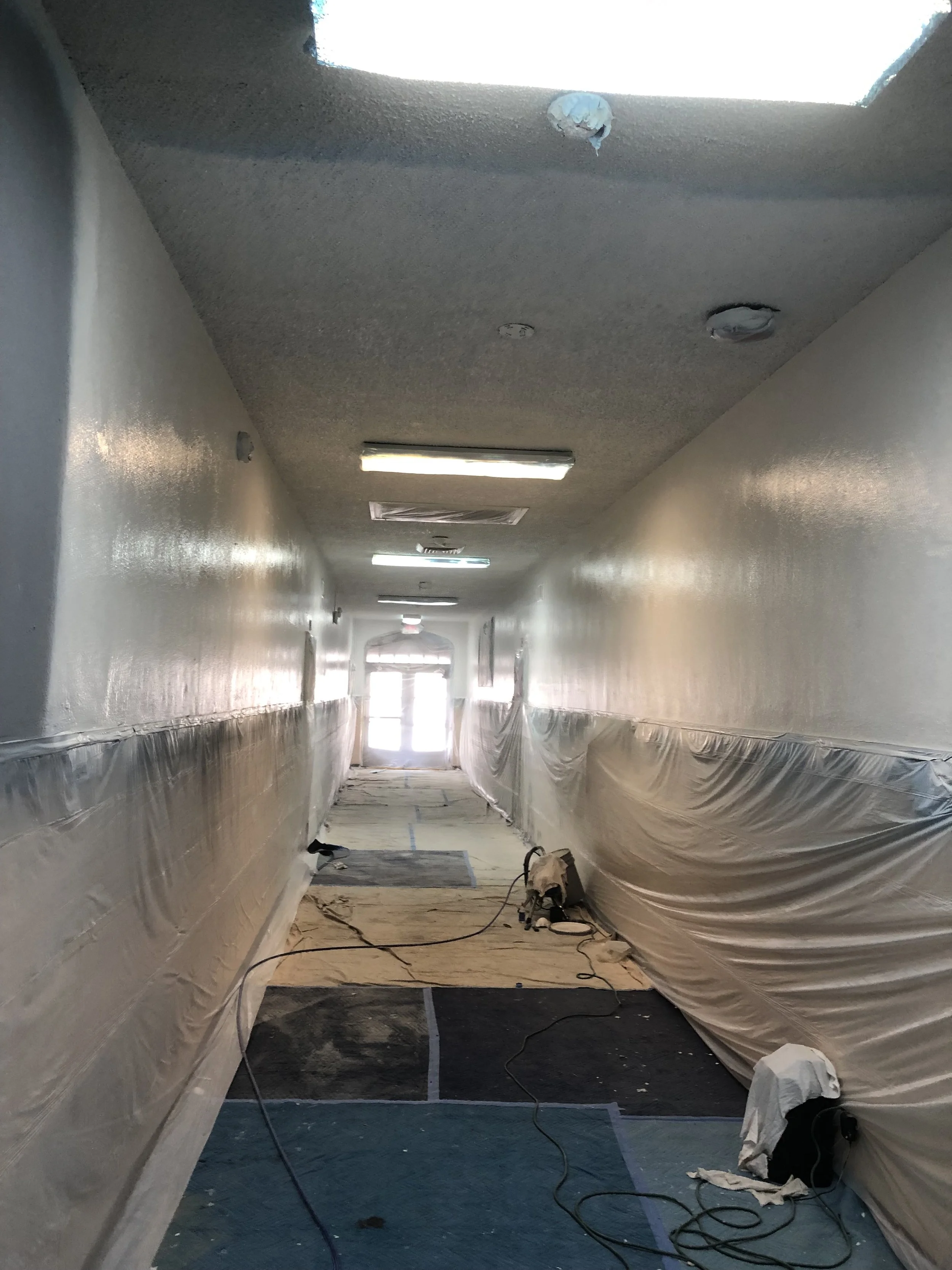 Long hallway under renovation with plastic sheeting on walls and floor mats, scattered tools and cables, overhead lighting, and a bright exit at the end.