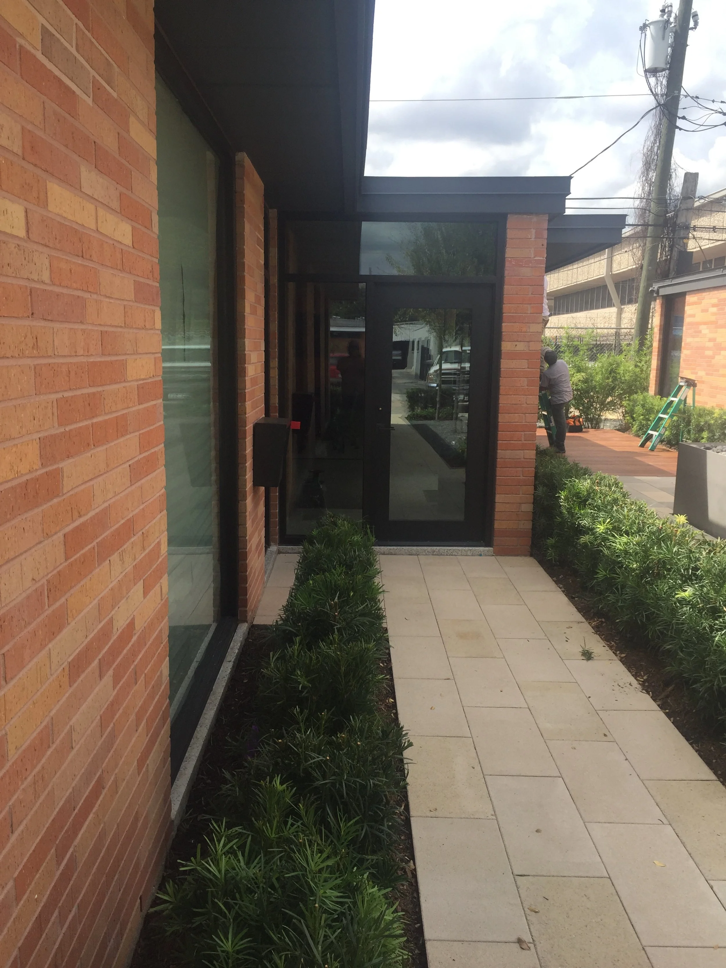 Brick building entrance with glass door, surrounded by green shrubs and paved walkway.