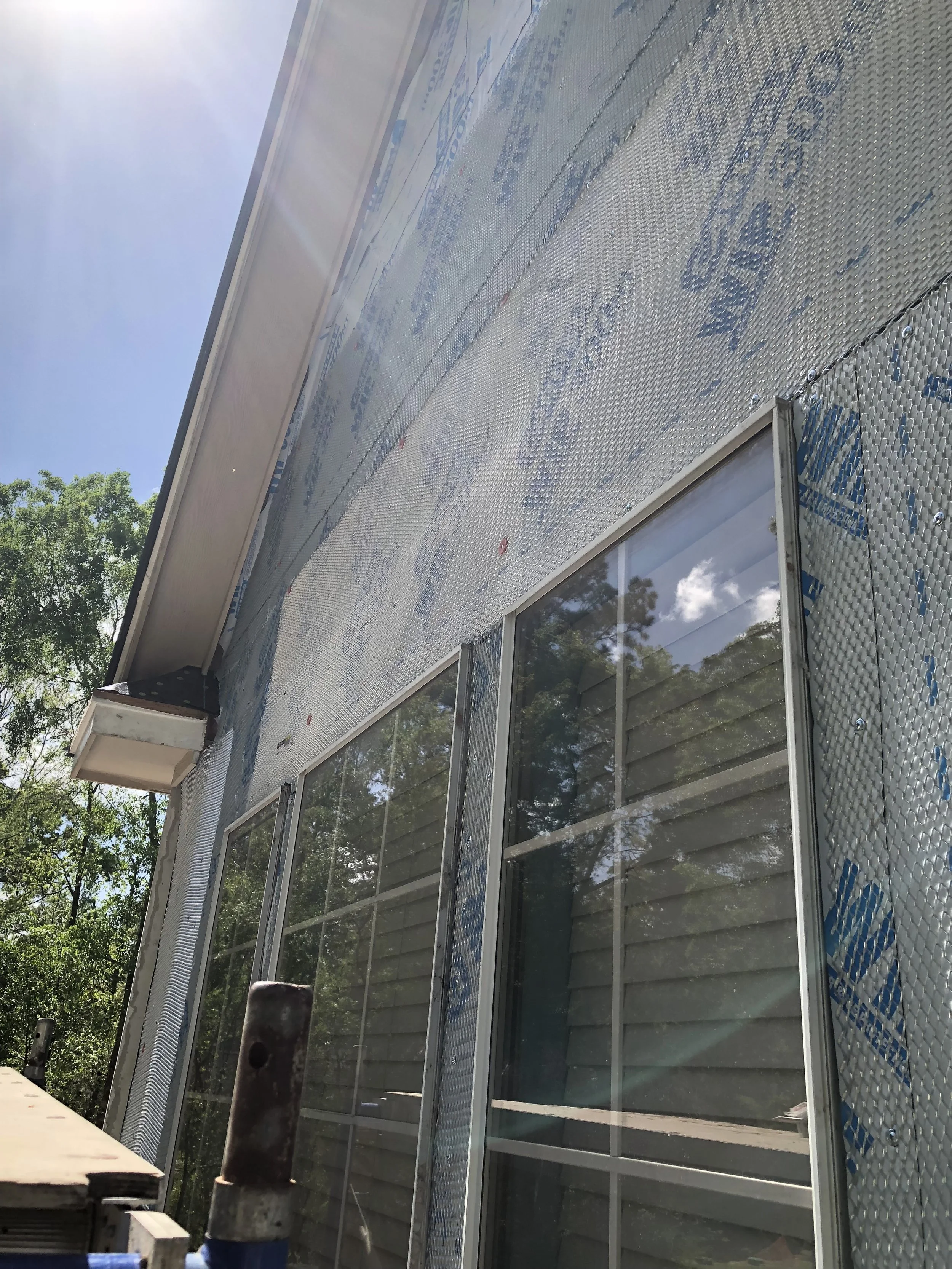 Wooden house wall under construction with weatherproof barrier, reflective insulation, and multiple windows against a clear sky.
