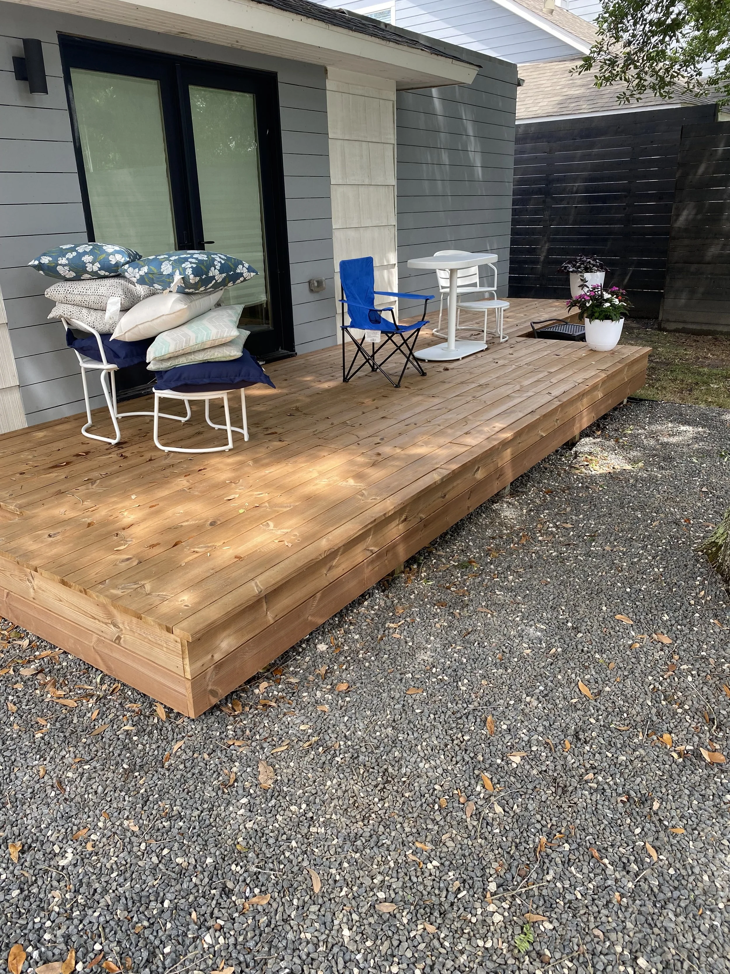 Wooden deck with outdoor furniture and cushions, including a blue folding chair and a small white table, next to a house with gray siding. A potted plant is on the deck and the ground is covered with gravel.