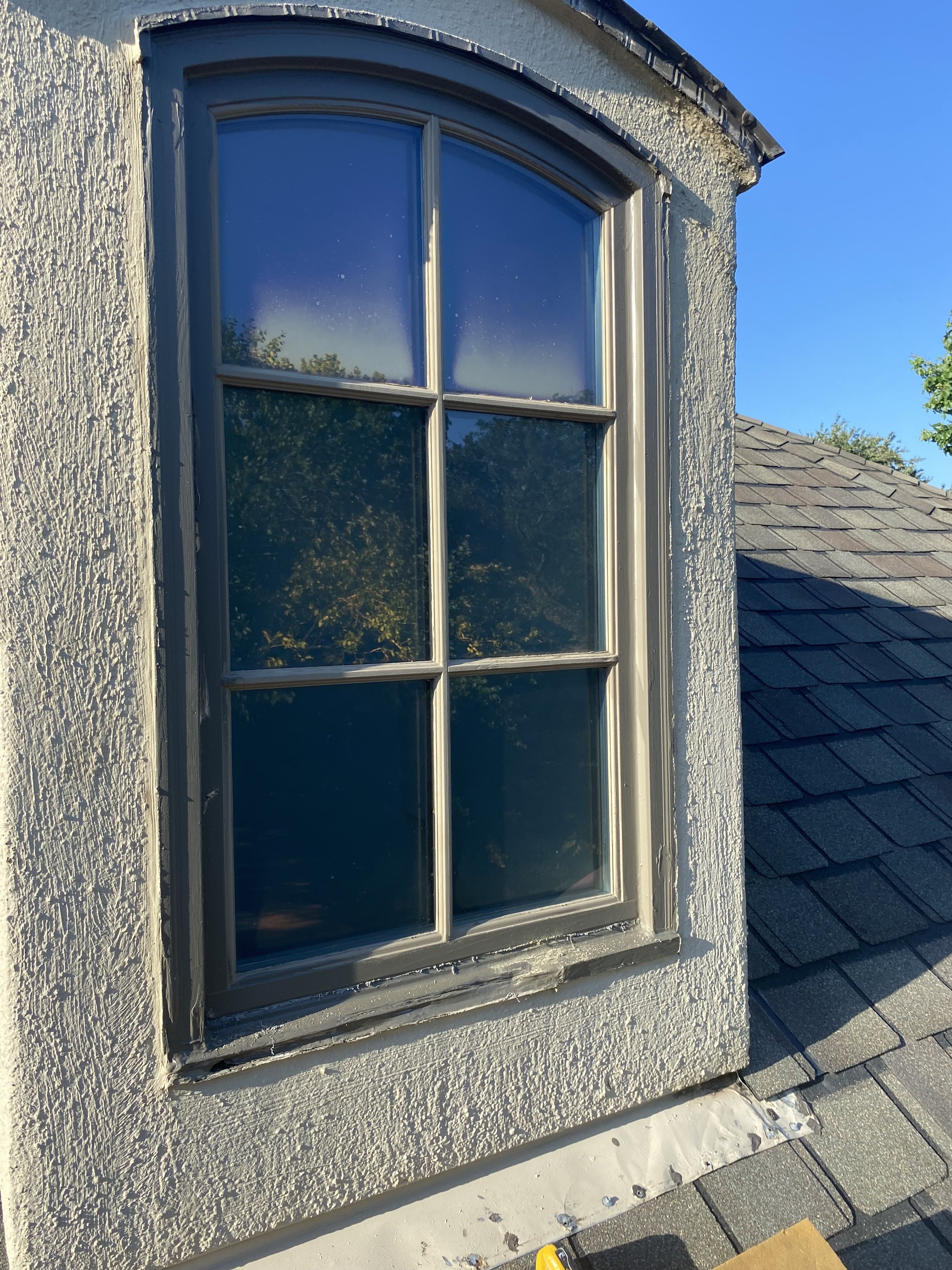 A rectangular window with four panes set in a textured wall, reflected trees and sky, above a shingled roof under blue sky.