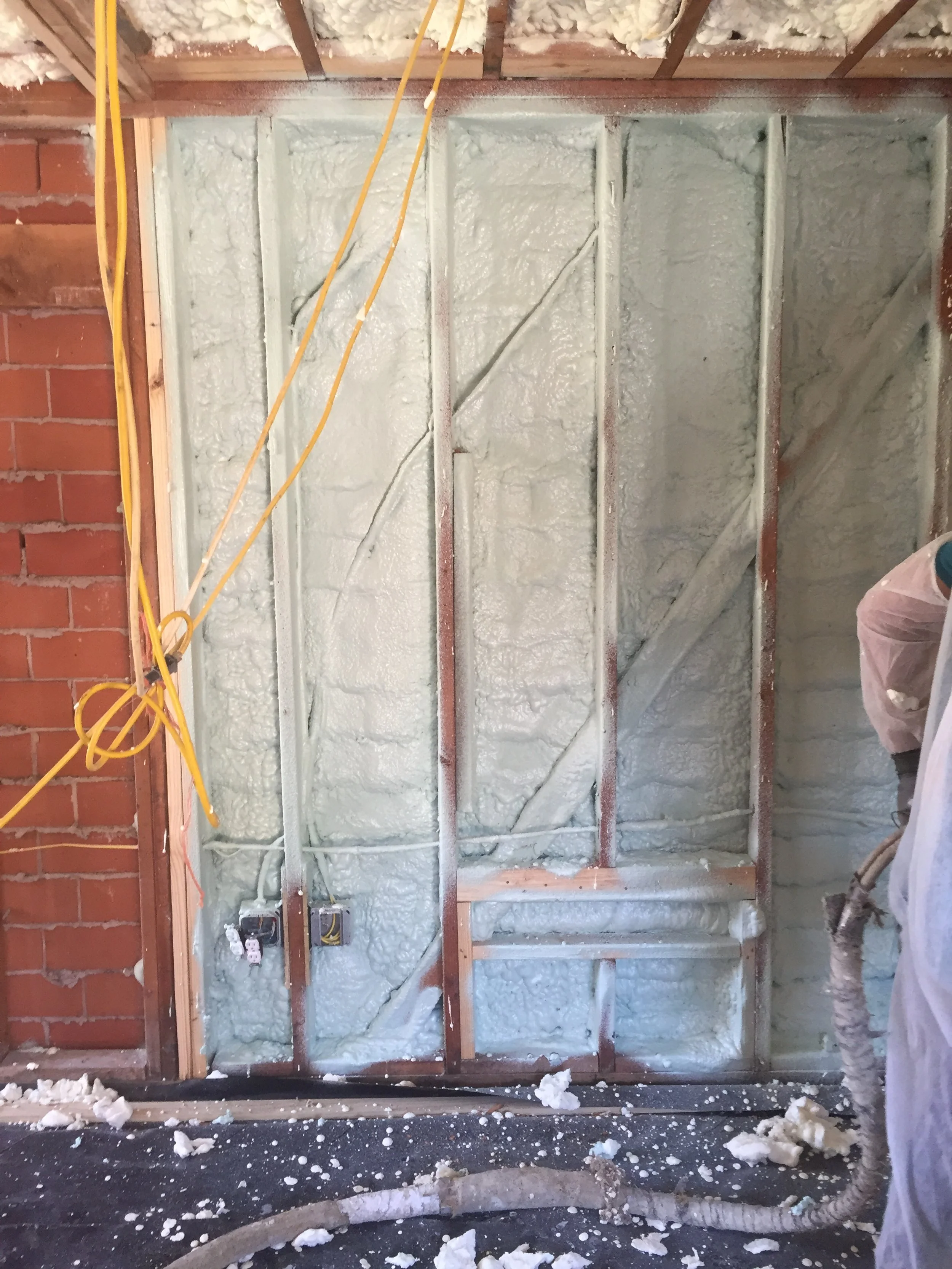Spray foam insulation applied to interior wall framing with exposed electrical wiring and outlets, construction site debris on the floor.