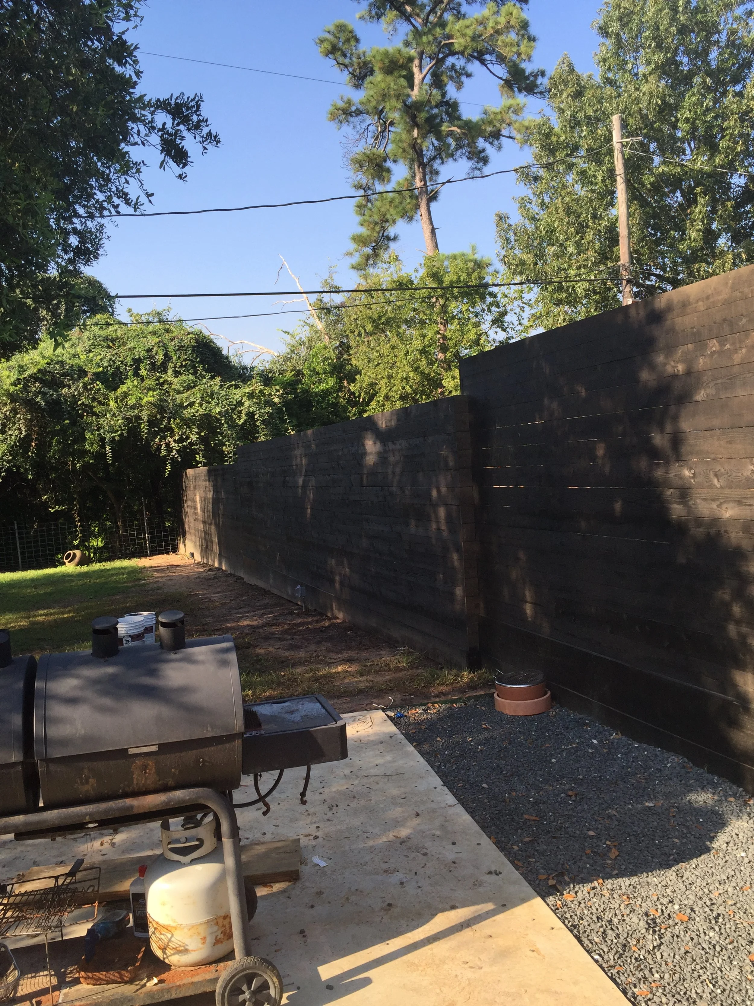 Backyard with a smoker grill, wooden fence, and trees under a blue sky.