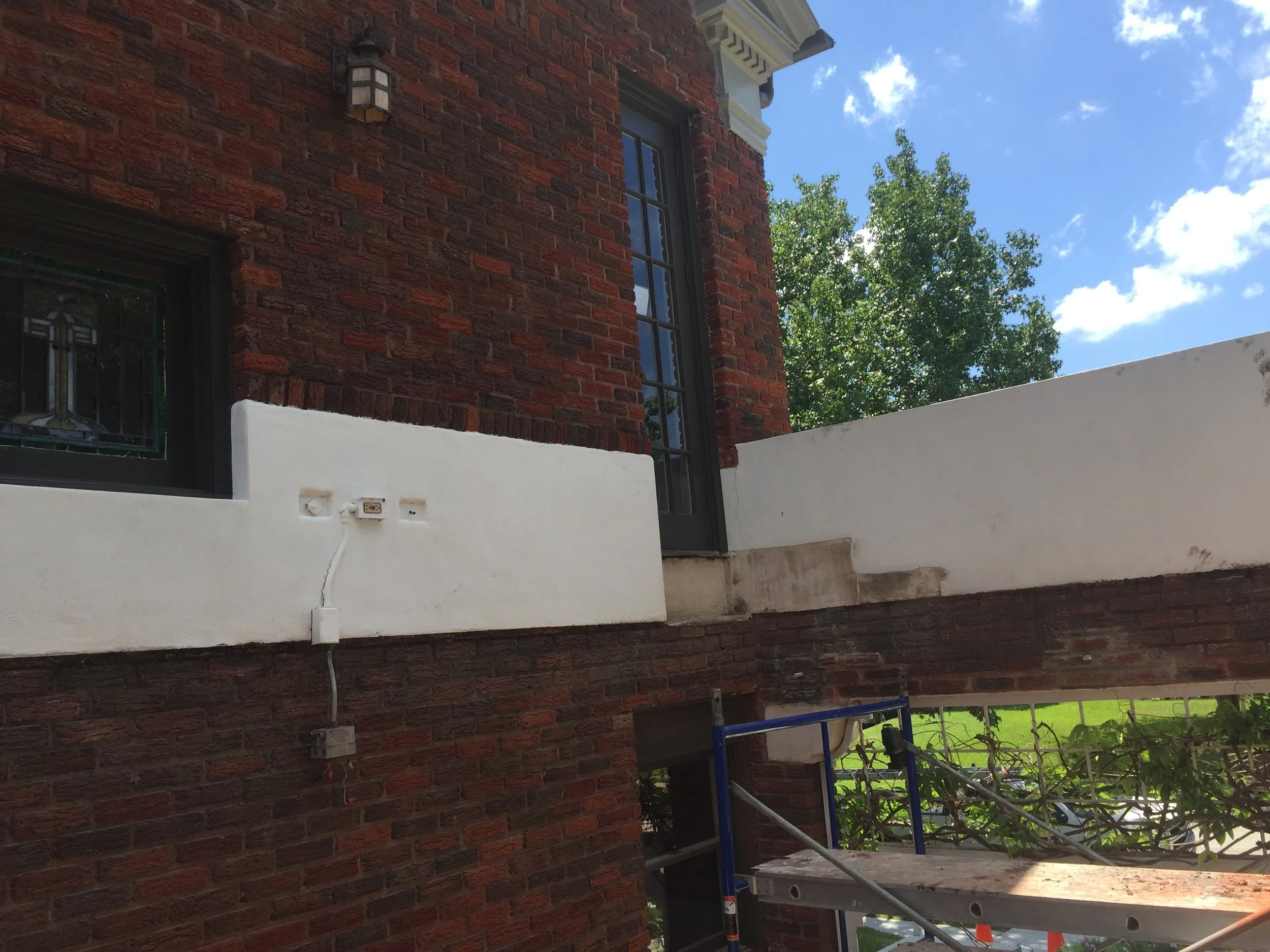 Red brick house with white stucco accents, outdoor light fixture, stained glass window, and a ladder. Trees and blue sky in the background.