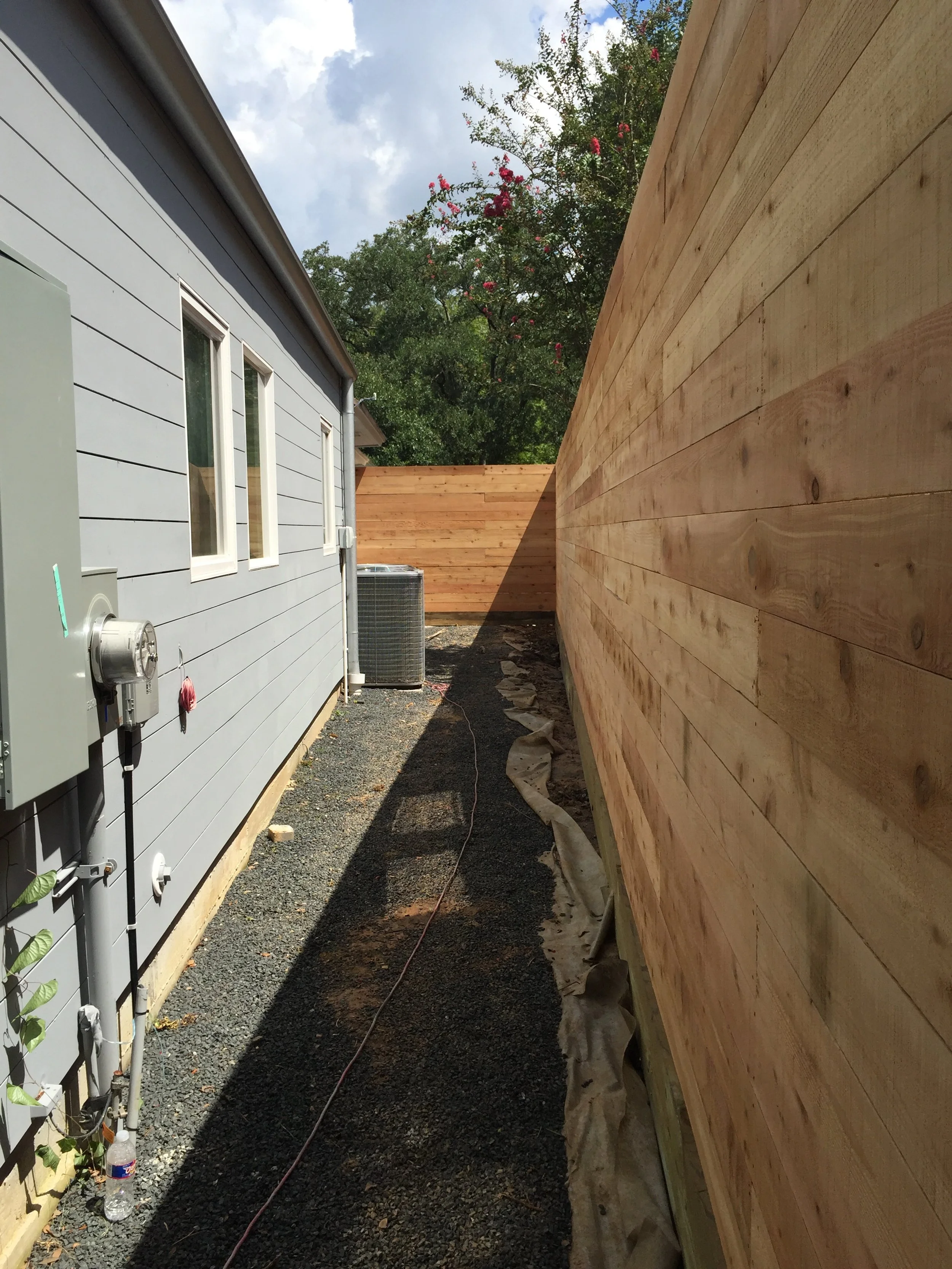 Narrow pathway between a house with blue siding and a wooden fence, featuring a gravel walkway and an air conditioning unit.