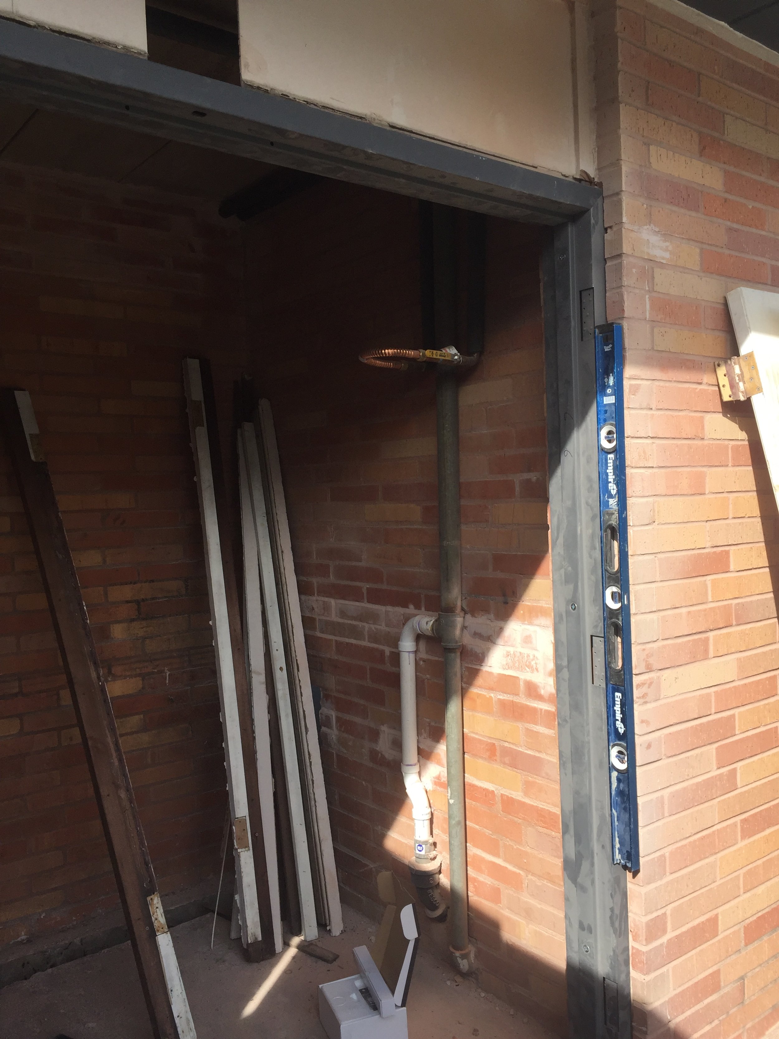 Red brick wall with construction tools, wooden planks, a blue level, and exposed pipes in a small space.