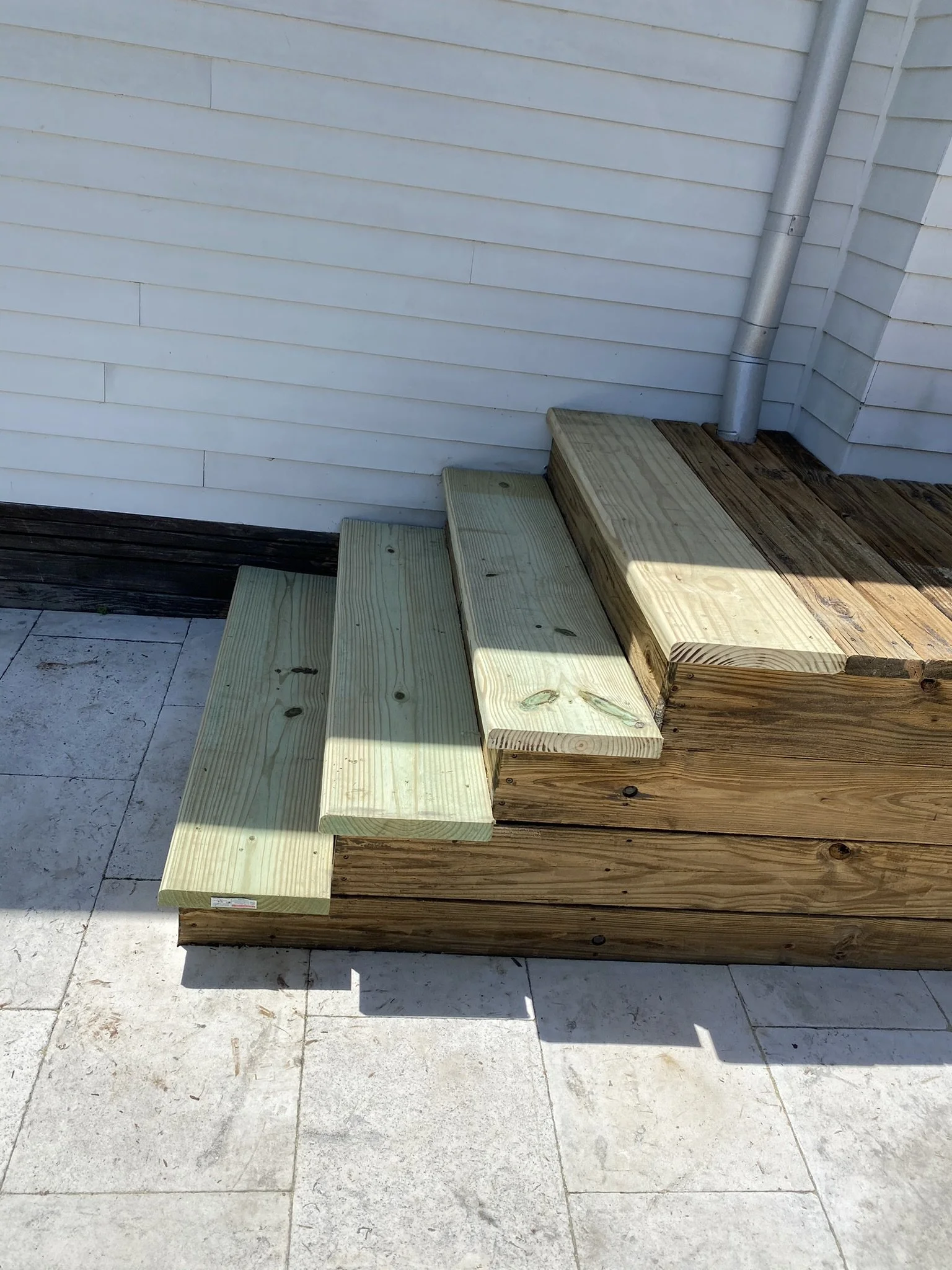 Wooden steps on a patio next to a white siding wall with a downspout.