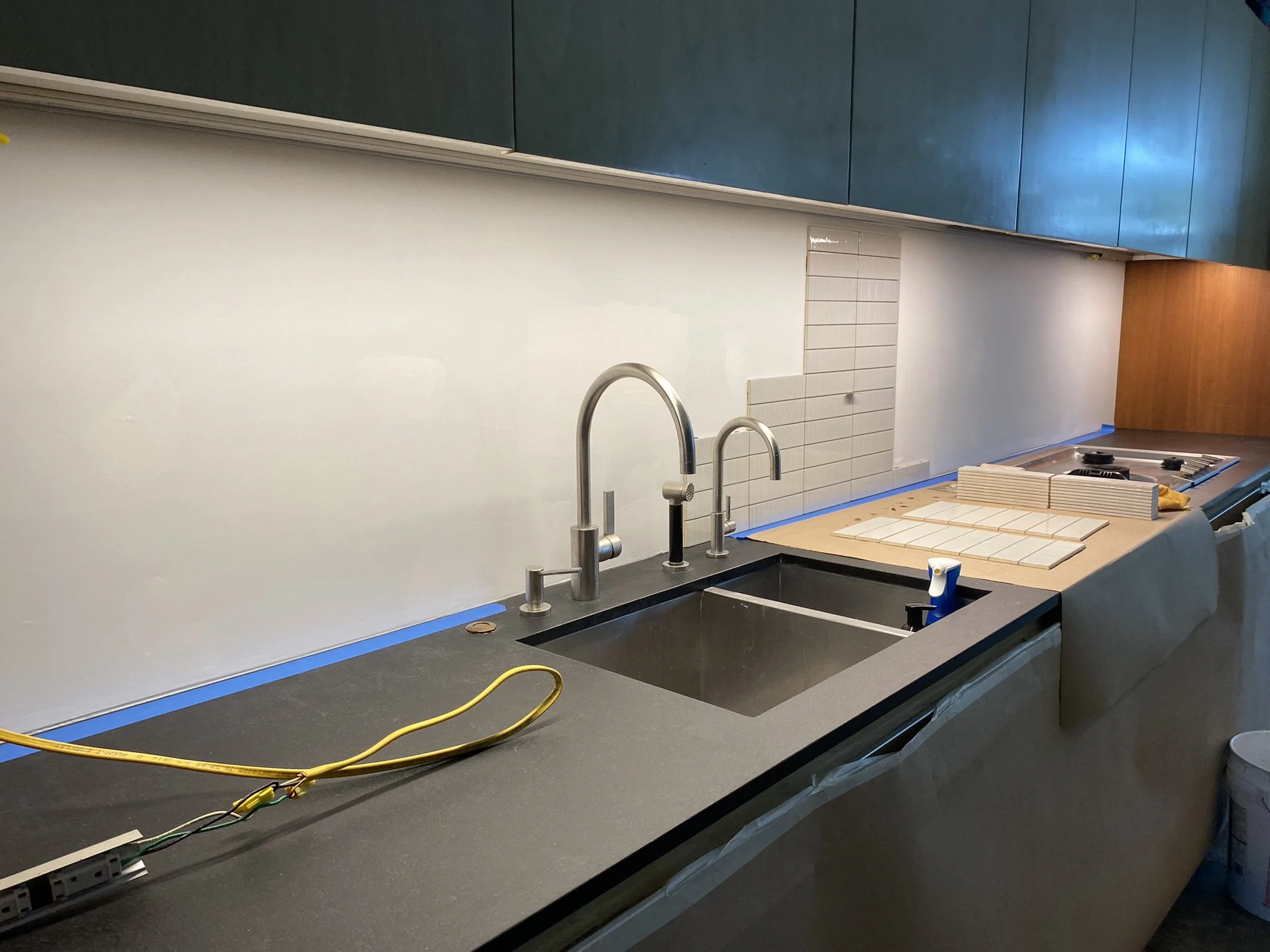 Modern kitchen with gray countertop, stainless steel sink and faucet, partial tile backsplash installation, overhead cabinets, and a stovetop on the right. Cables and tools indicate ongoing renovation.