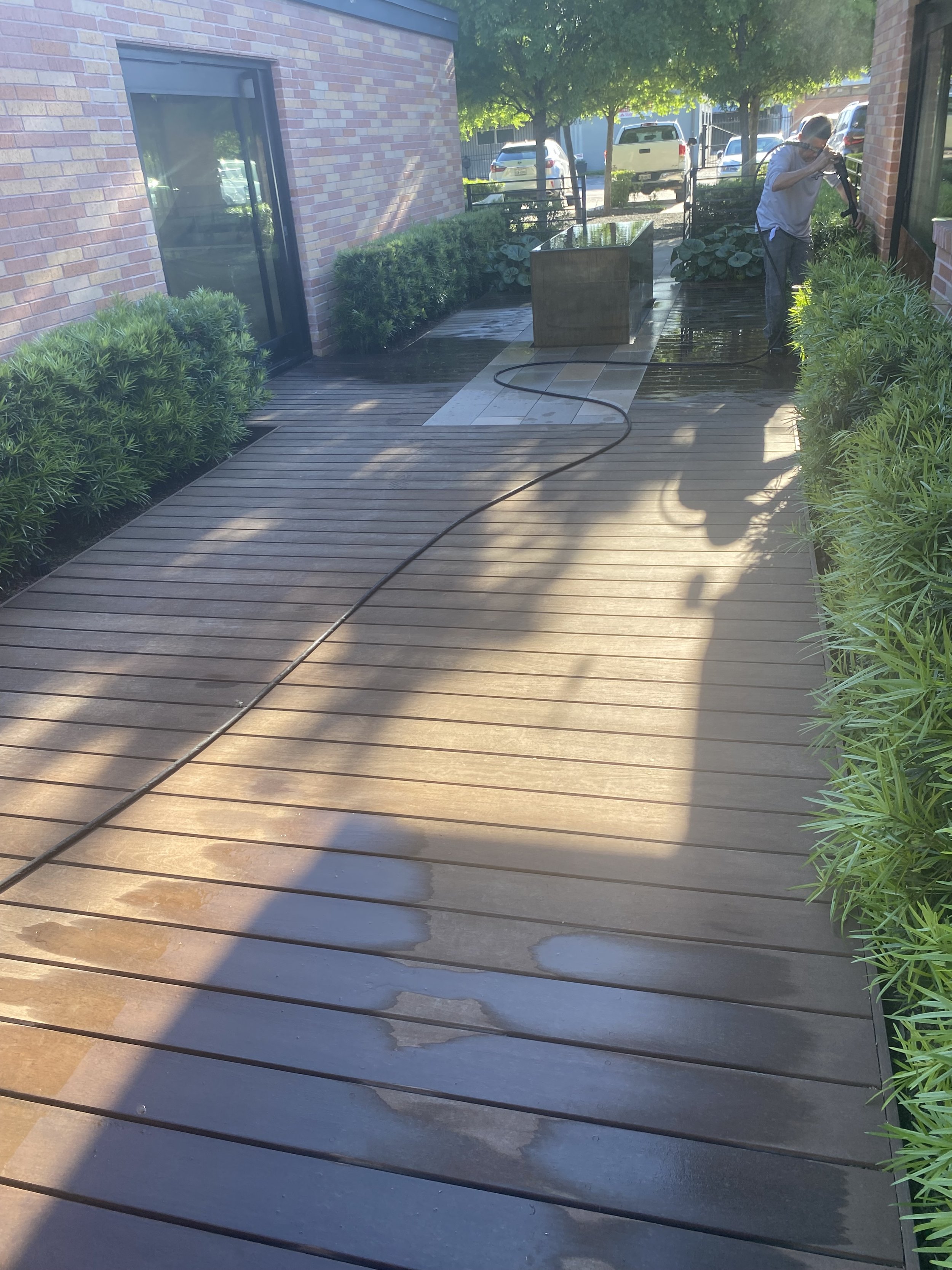 Wooden deck being pressure washed near brick building and plants, with a person cleaning in the background.