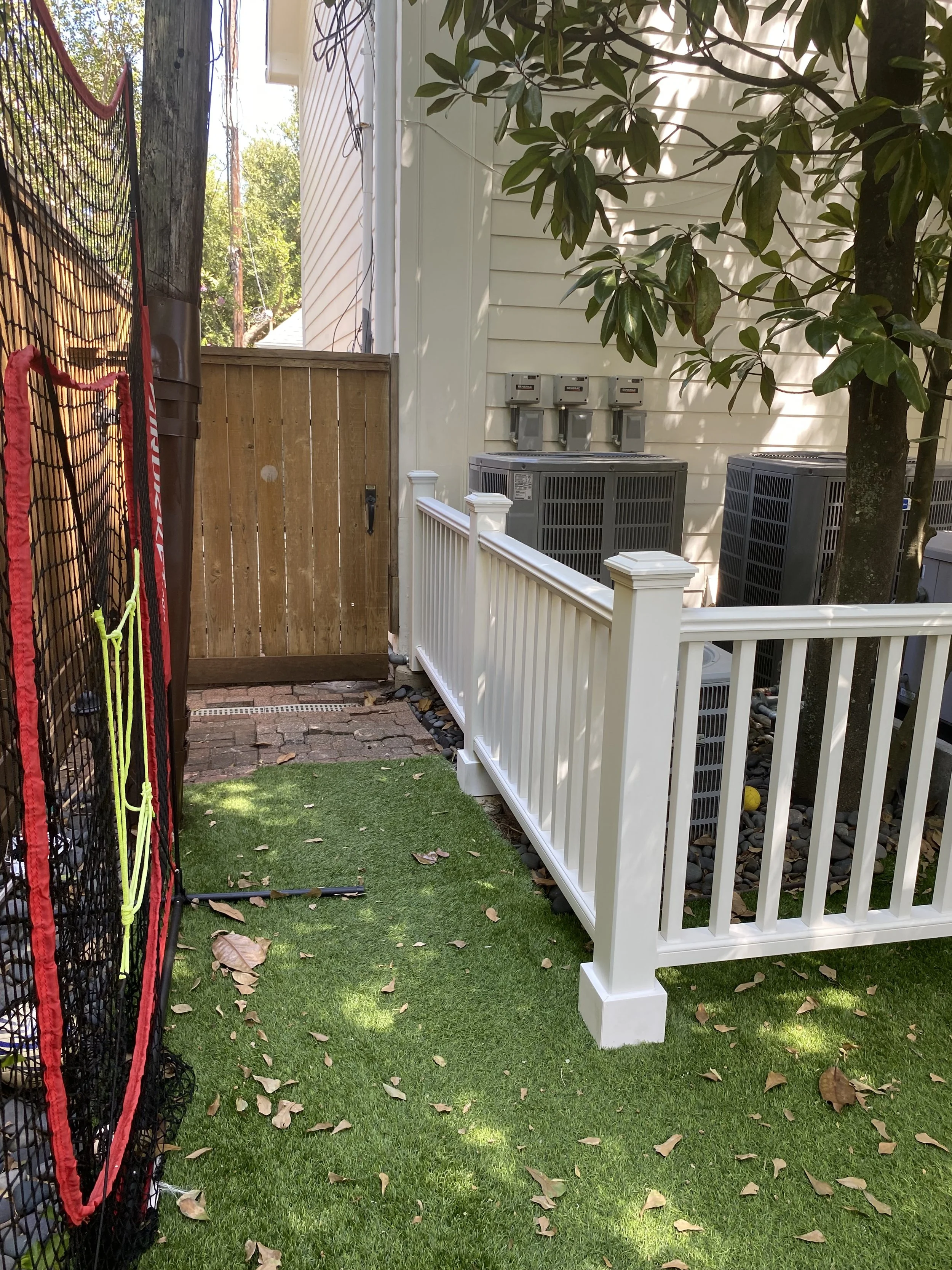 Side yard with artificial grass, a black and red sports net, white railing, wooden gate, air conditioning units, and a tree with green leaves.