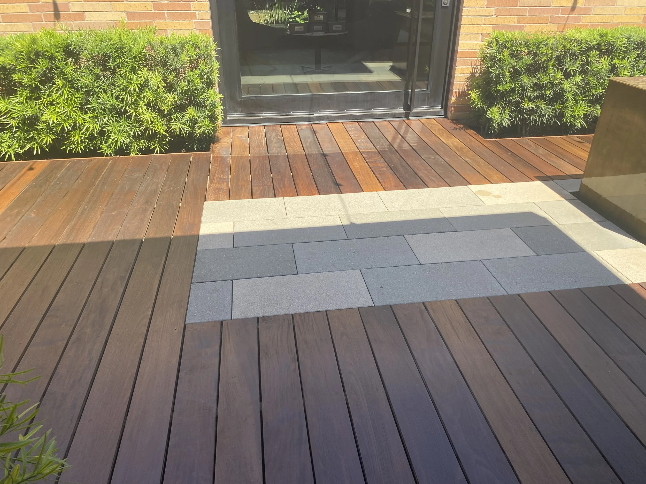 Patio with wooden deck and pavers, surrounded by greenery and brick wall