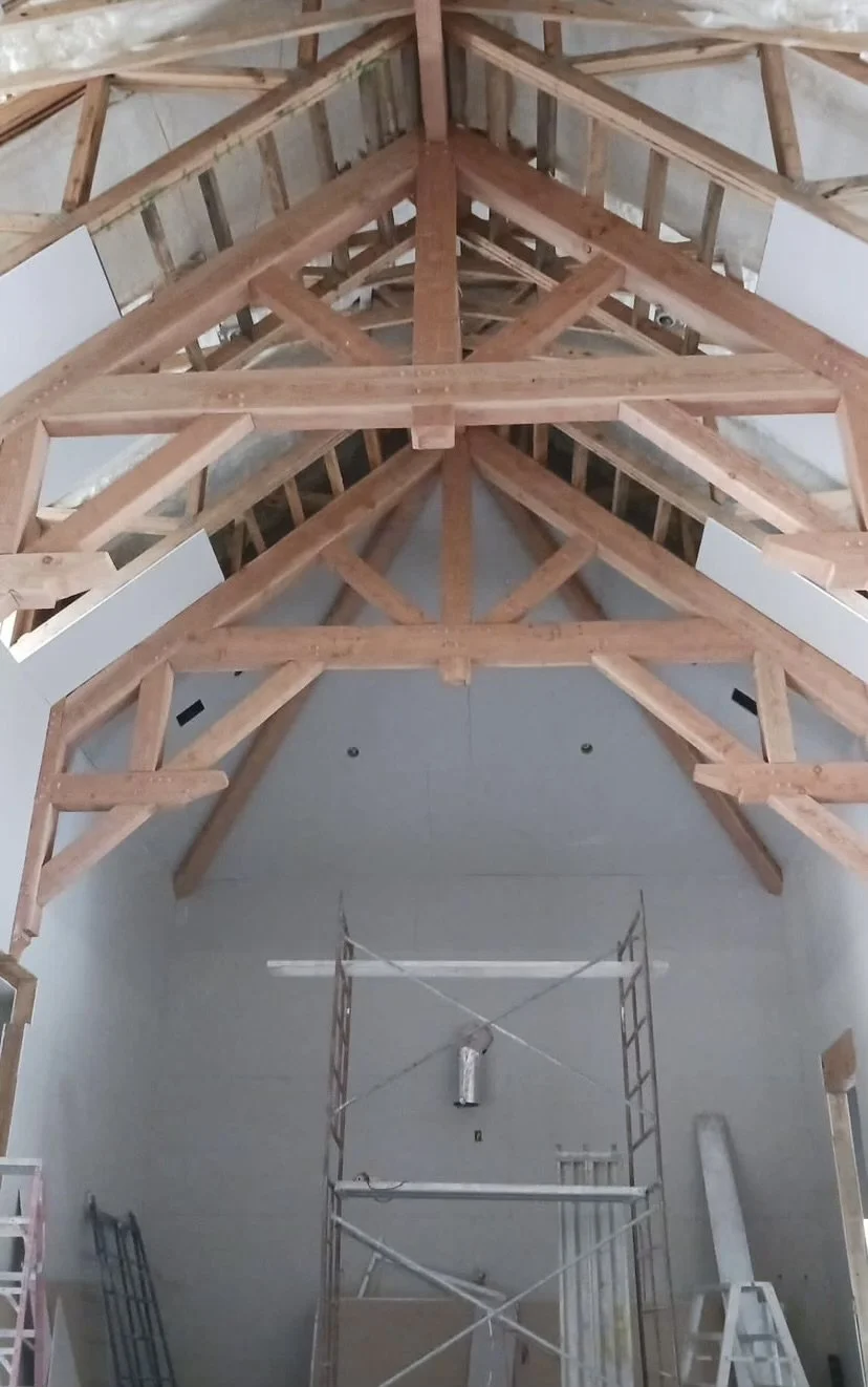 Interior view of a building under construction with exposed wooden roof trusses and scaffolding.