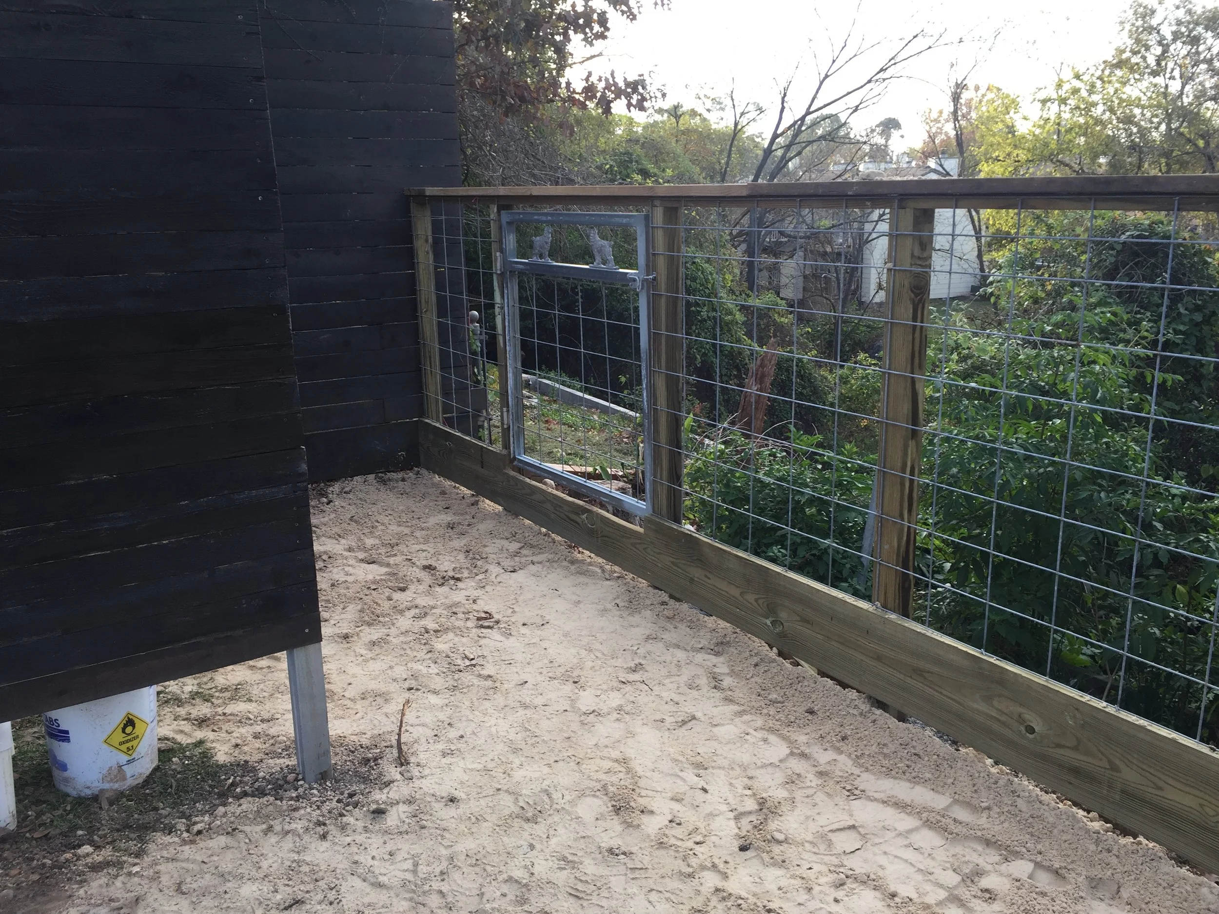 Outdoor area with a partially constructed wooden and wire fence, black painted wall, and sandy ground.