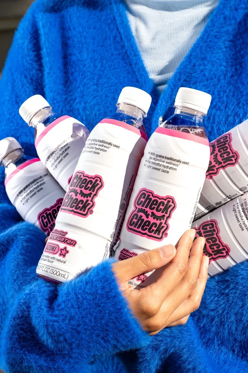 A person wearing a bright blue fuzzy coat holding several bottles of CheckCheck rice ater with white and pink labels.