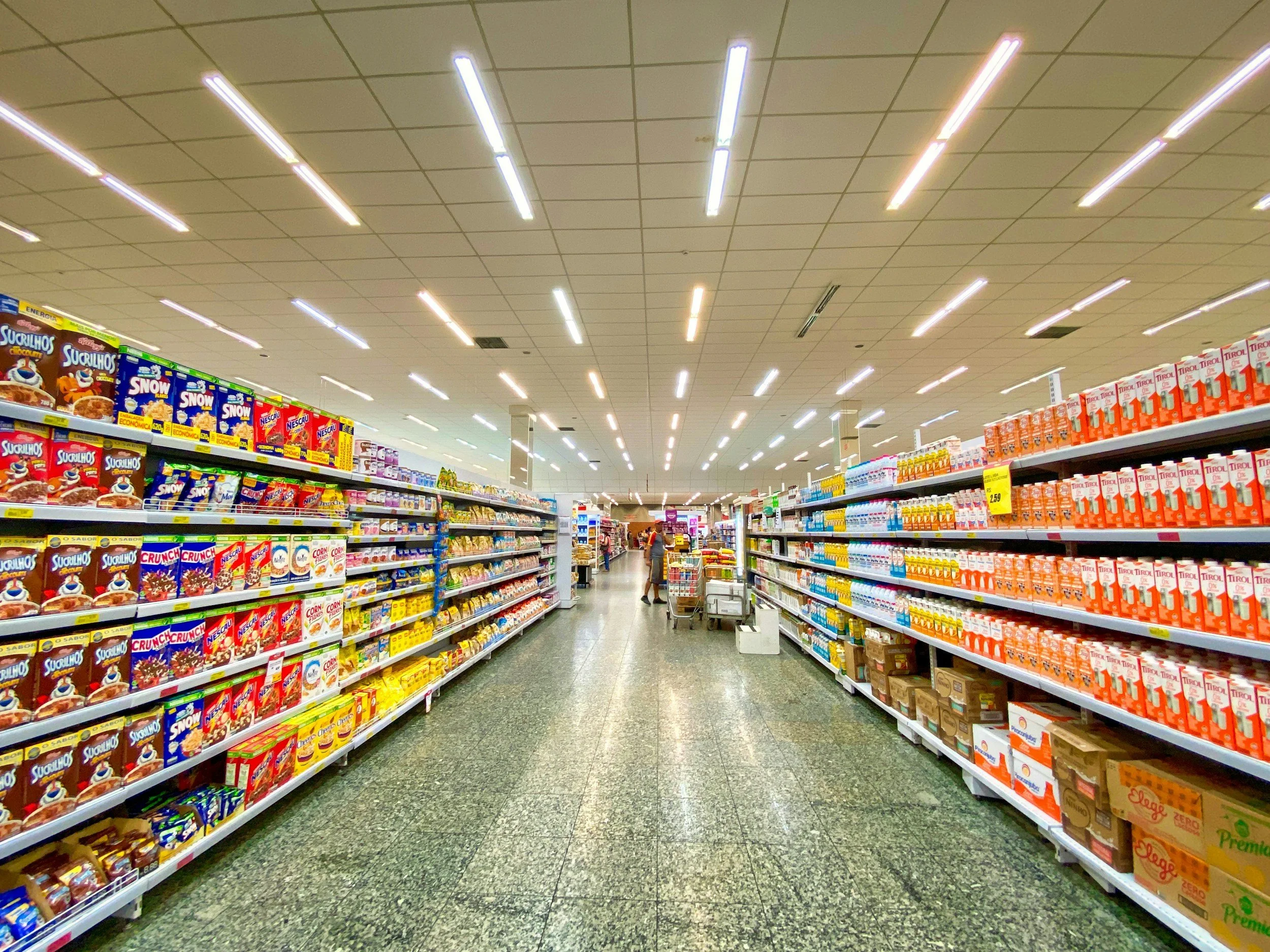 A grocery store aisle with snack and beverage shelves, fluorescent lighting overhead, and a shopper pushing a cart in the distance.