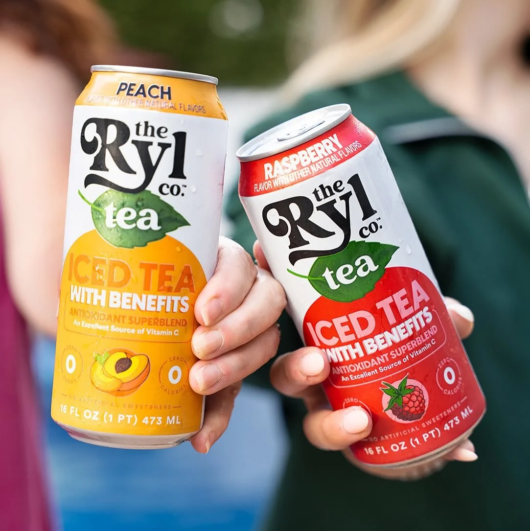 Two hands holding cans of The RYL Co. iced tea, one peach flavored and one raspberry flavored, with a blurred person in the background.
