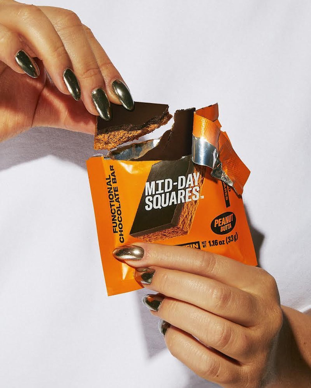 Close-up of a person with metallic nail polish holding a partially opened package of Mid-Day Squares peanut butter chocolate protein bar, with a piece of the bar being lifted out.
